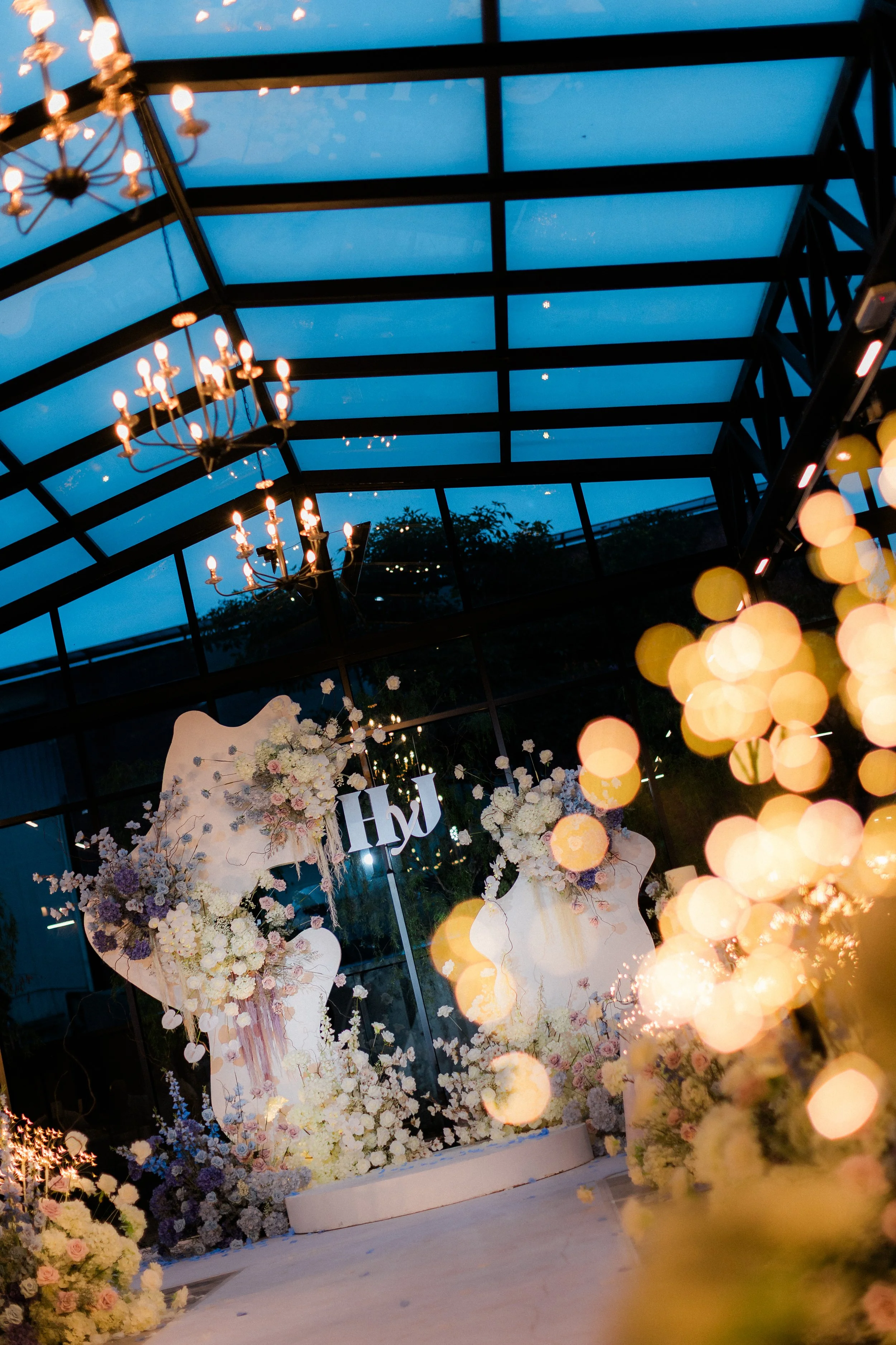 Indoor wedding venue decorated with white and pink flowers, illuminated by chandeliers, with a large sign displaying initials 'H' and 'J' in the background.