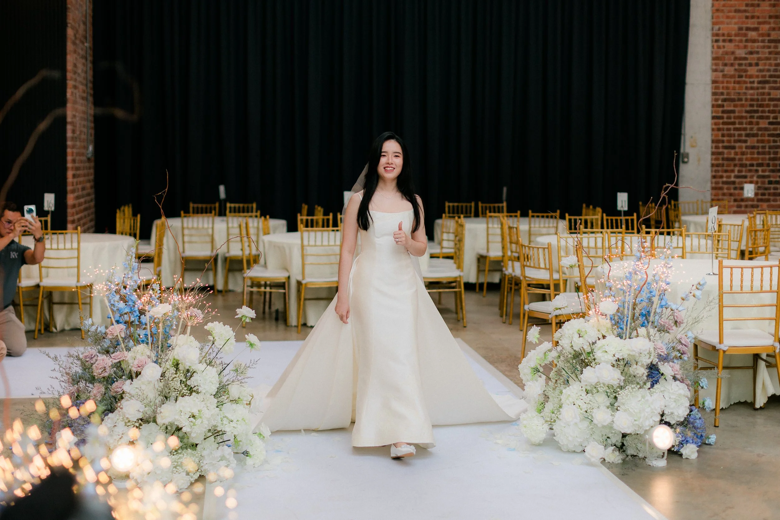 A woman in a white wedding dress giving a thumbs up and smiling at a wedding reception, with floral arrangements and guest seating around her.