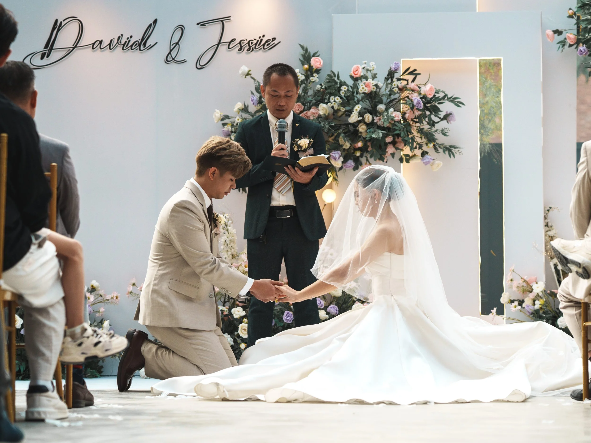 A wedding ceremony with a bride in a white gown and veil, and a groom in a beige suit holding hands with their eyes closed, kneeling on a stage decorated with flowers. An officiant stands behind them reading from a book. Guests are seated on chairs.