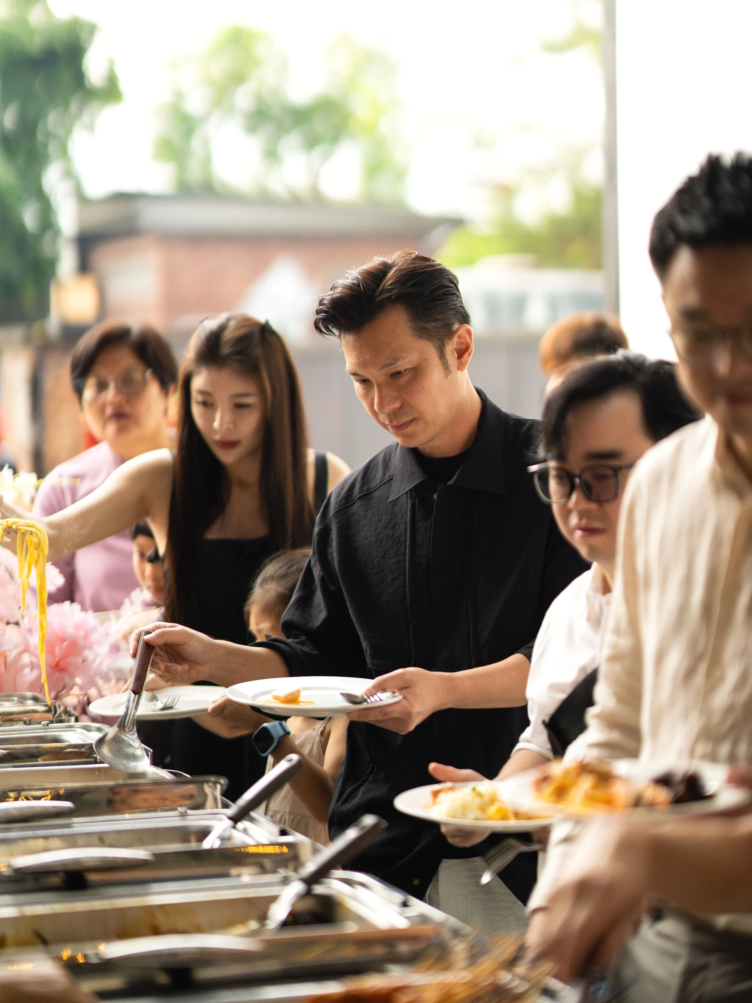 People serving themselves food from a buffet, holding plates and serving utensils at an outdoor event.