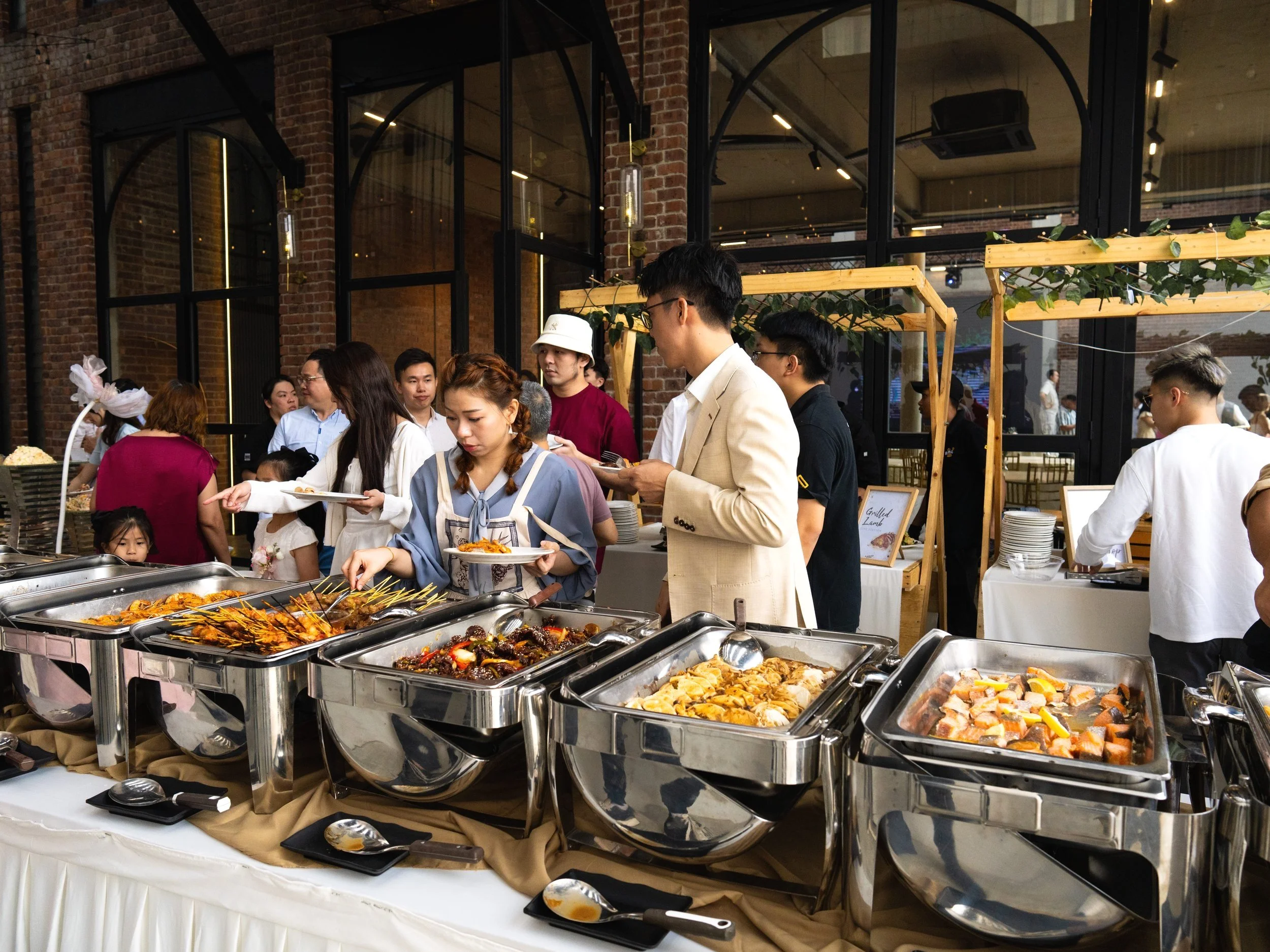 People serving themselves food at a buffet in a modern, stylish venue with brick walls and large glass windows.