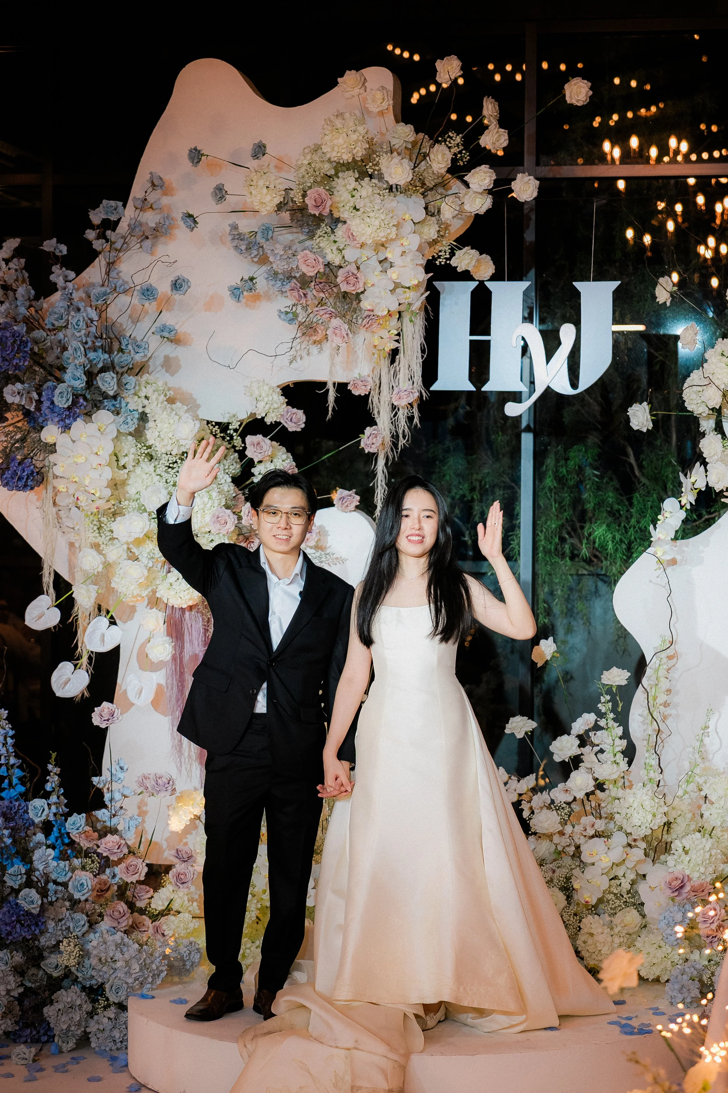 A couple dressed in formal wedding attire standing on a decorated platform, waving at the camera, with floral arrangements and a large decorative sign behind them.