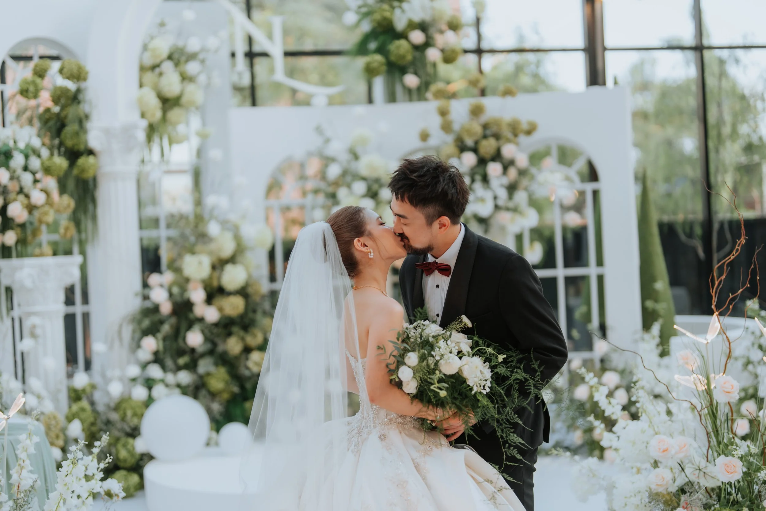A bride and groom sharing a kiss during their wedding ceremony in an elegant indoor venue decorated with white and green floral arrangements.