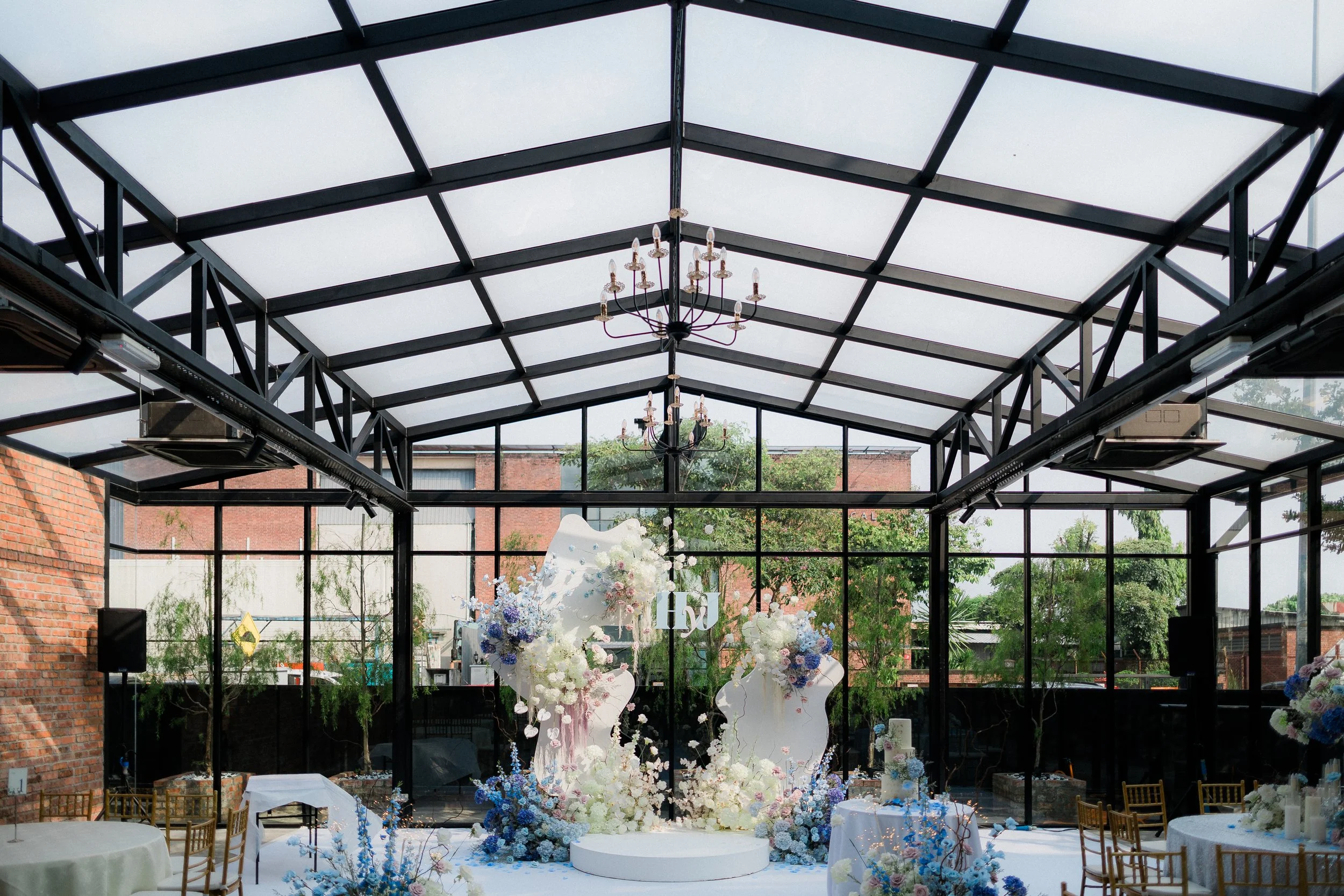 Indoor wedding reception area with large flower arrangements in pastel colors, white platform, and a glass ceiling with chandeliers, outside trees visible through glass walls.