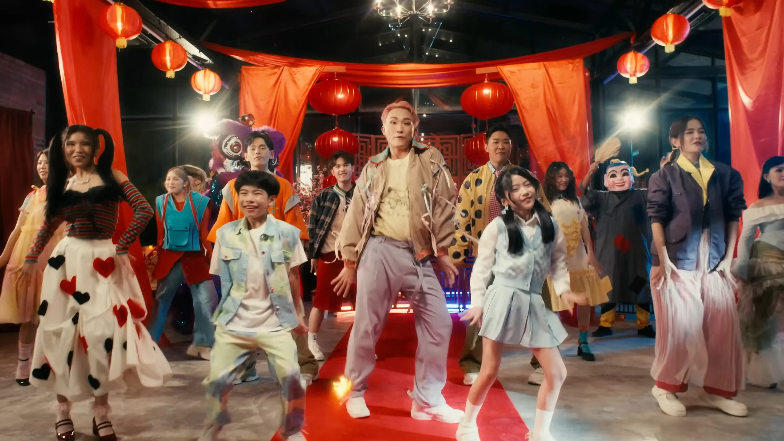 A diverse group of people, including children and adults, are performing a dance on a decorated stage with red lanterns and orange curtains, celebrating a festive event.