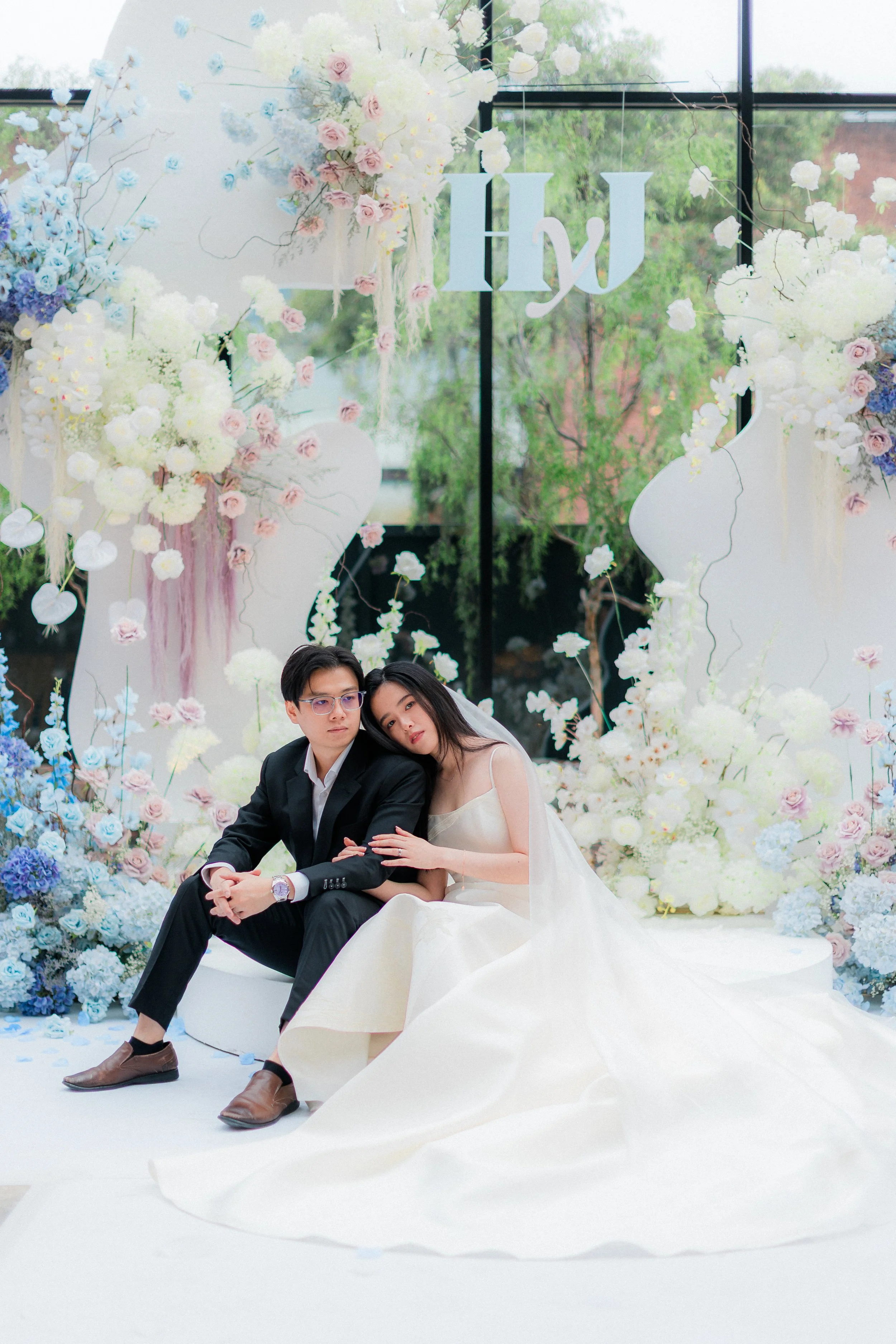 A bride and groom sitting together at a wedding, surrounded by white and pastel-colored flowers, with a decorative backdrop behind them.