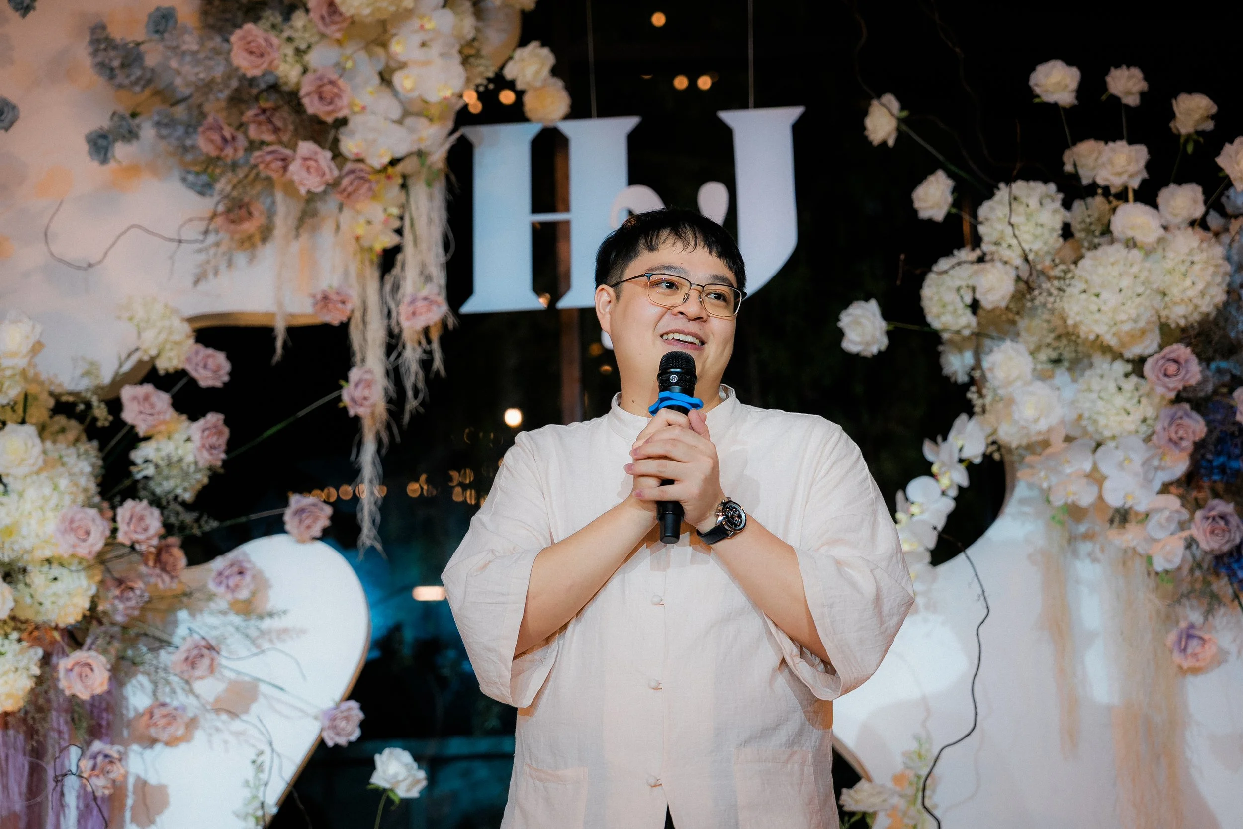 A man holding a microphone, standing in front of a floral wedding arch with the letters 'H' and 'J' in the background.