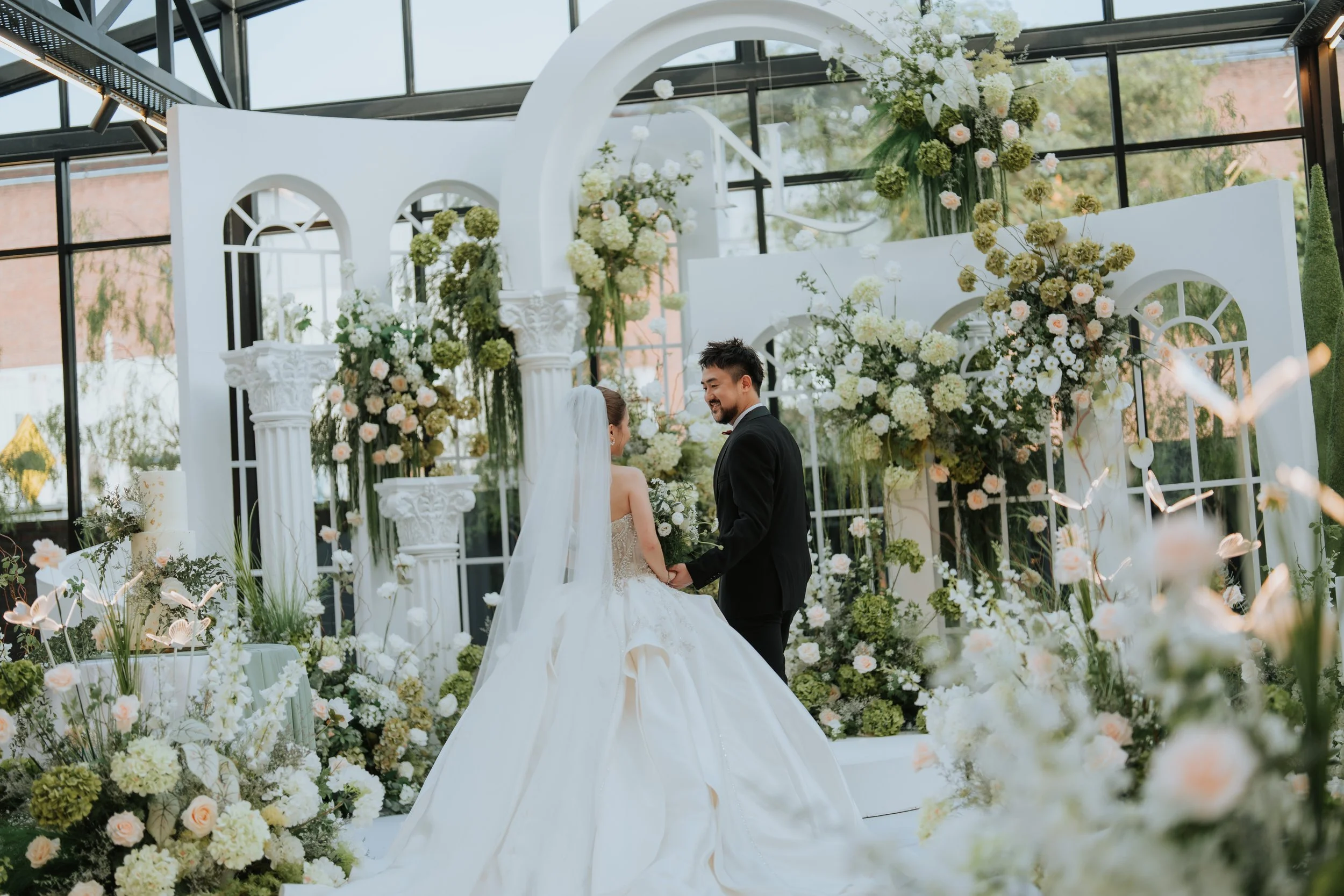 A bride and groom holding hands in a decorated wedding venue with white flowers and greenery, under an arched structure with large windows.