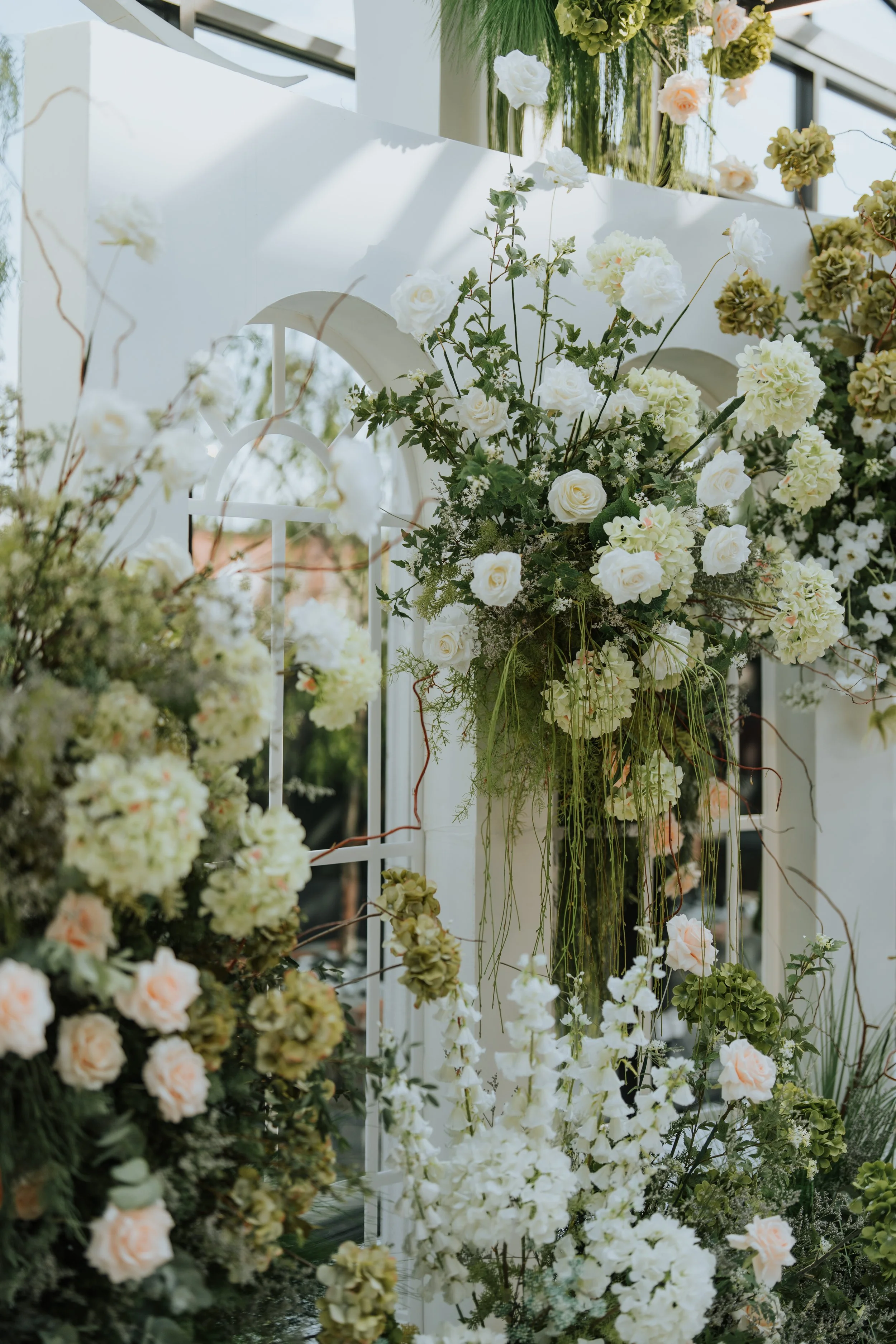 A floral arrangement featuring white roses, hydrangeas, and greenery in a garden setting, with a white trellis and a building with glass windows in the background.