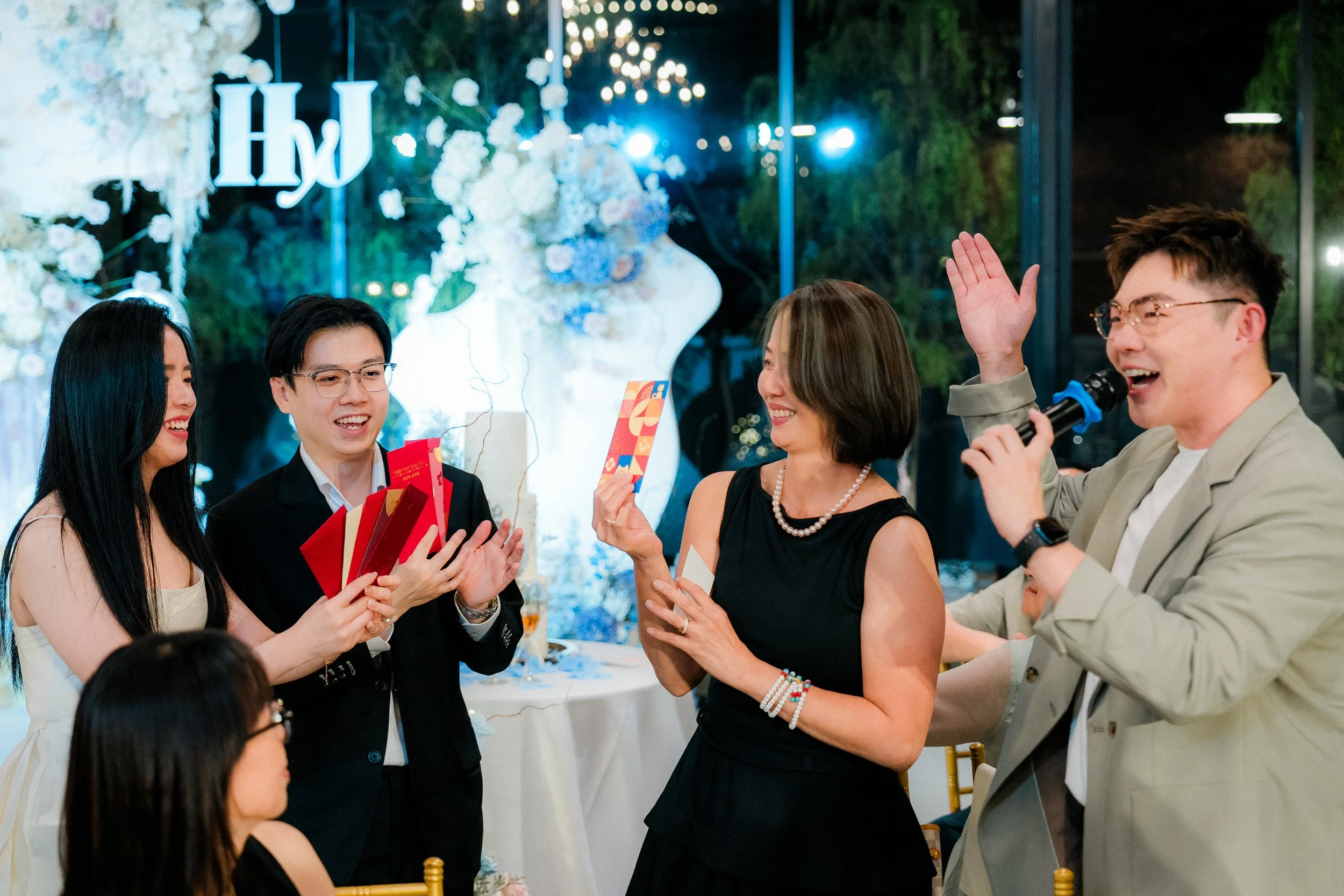 A group of five people celebrating at a decorated event, with a woman in a black dress receiving a red envelope, others clapping and smiling, a man speaking into a microphone, and floral decorations in the background.