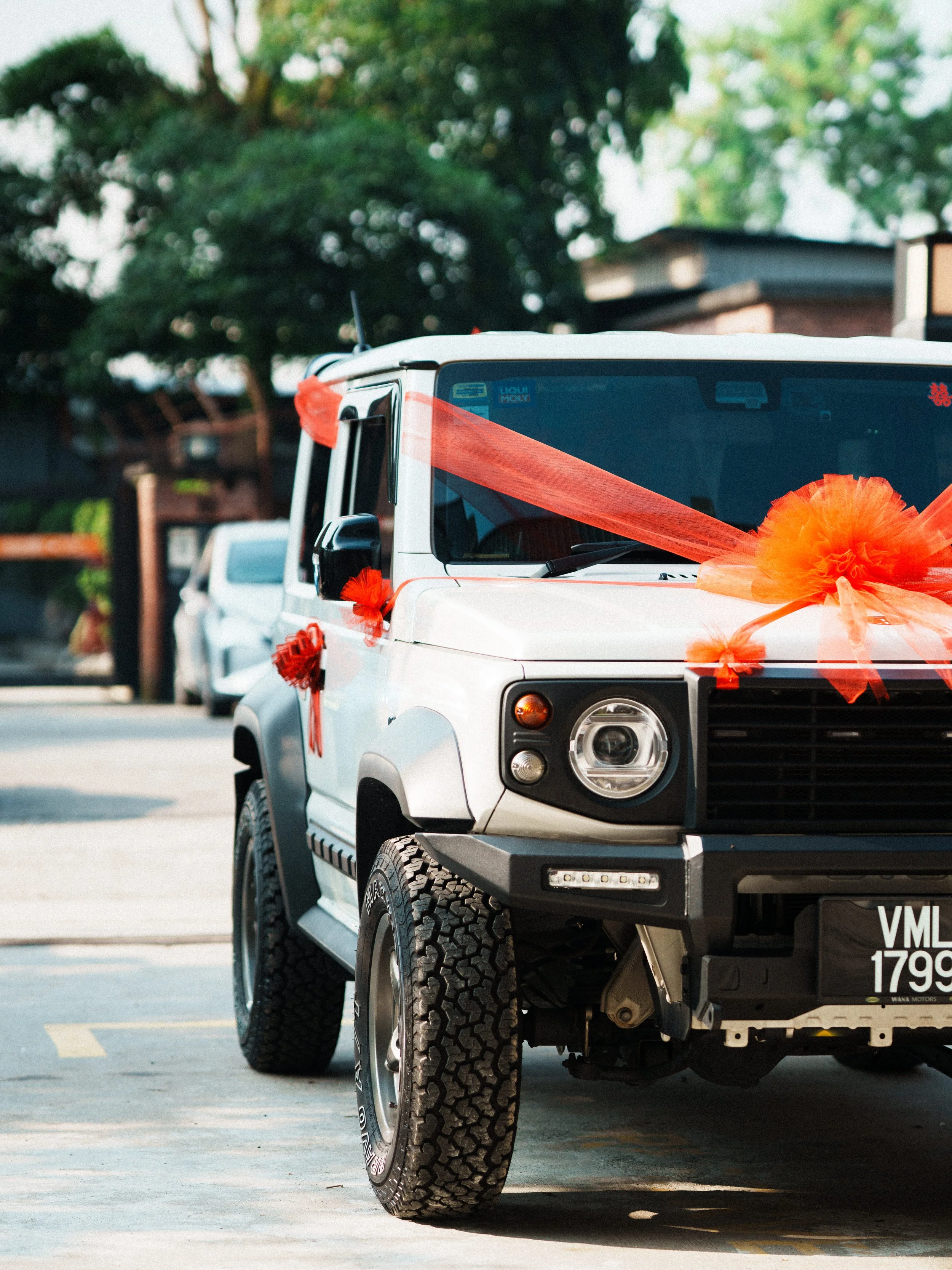 White off-road vehicle decorated with orange ribbons and bows for a celebration, parked outdoors with trees and other vehicles in the background.