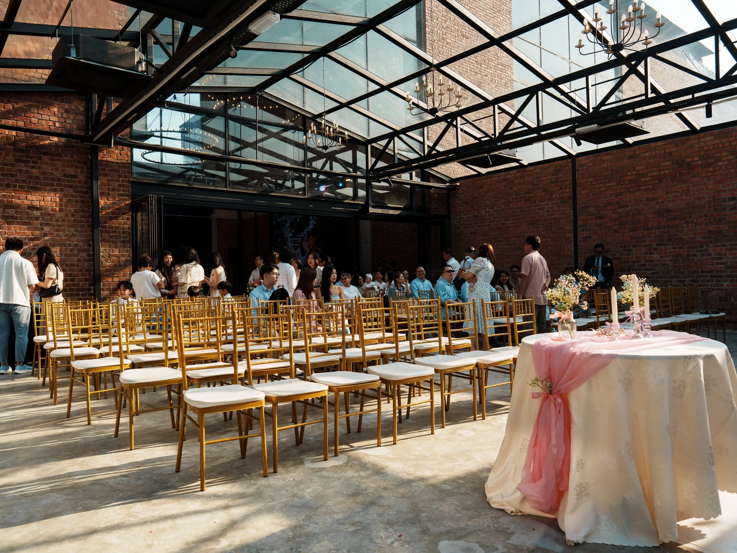 People attending a wedding or event in a partially outdoor space with glass roof and brick walls, with rows of golden chairs, a table with pink fabric and floral arrangements, and some guests standing and sitting.