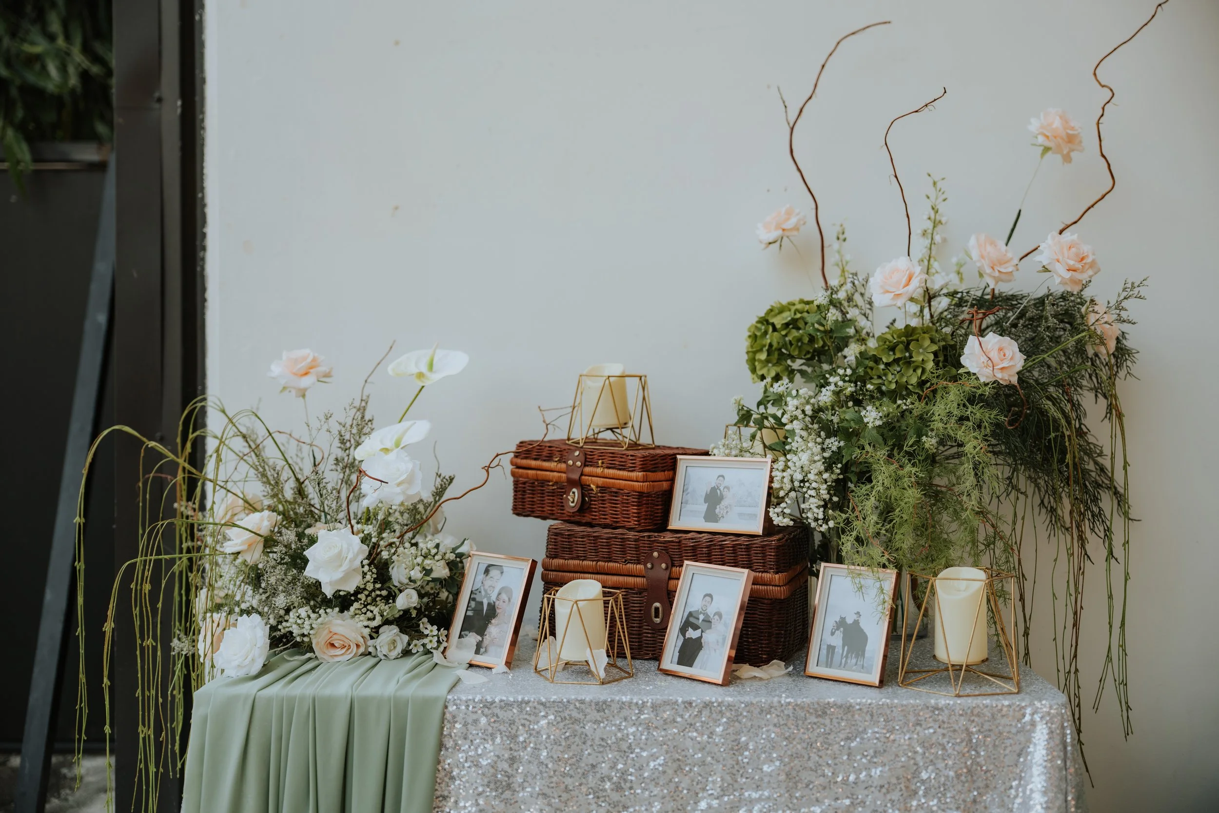 A decorative display with floral arrangements, framed photographs, candles, and wicker baskets on a sparkling tablecloth, likely at a wedding or special event.