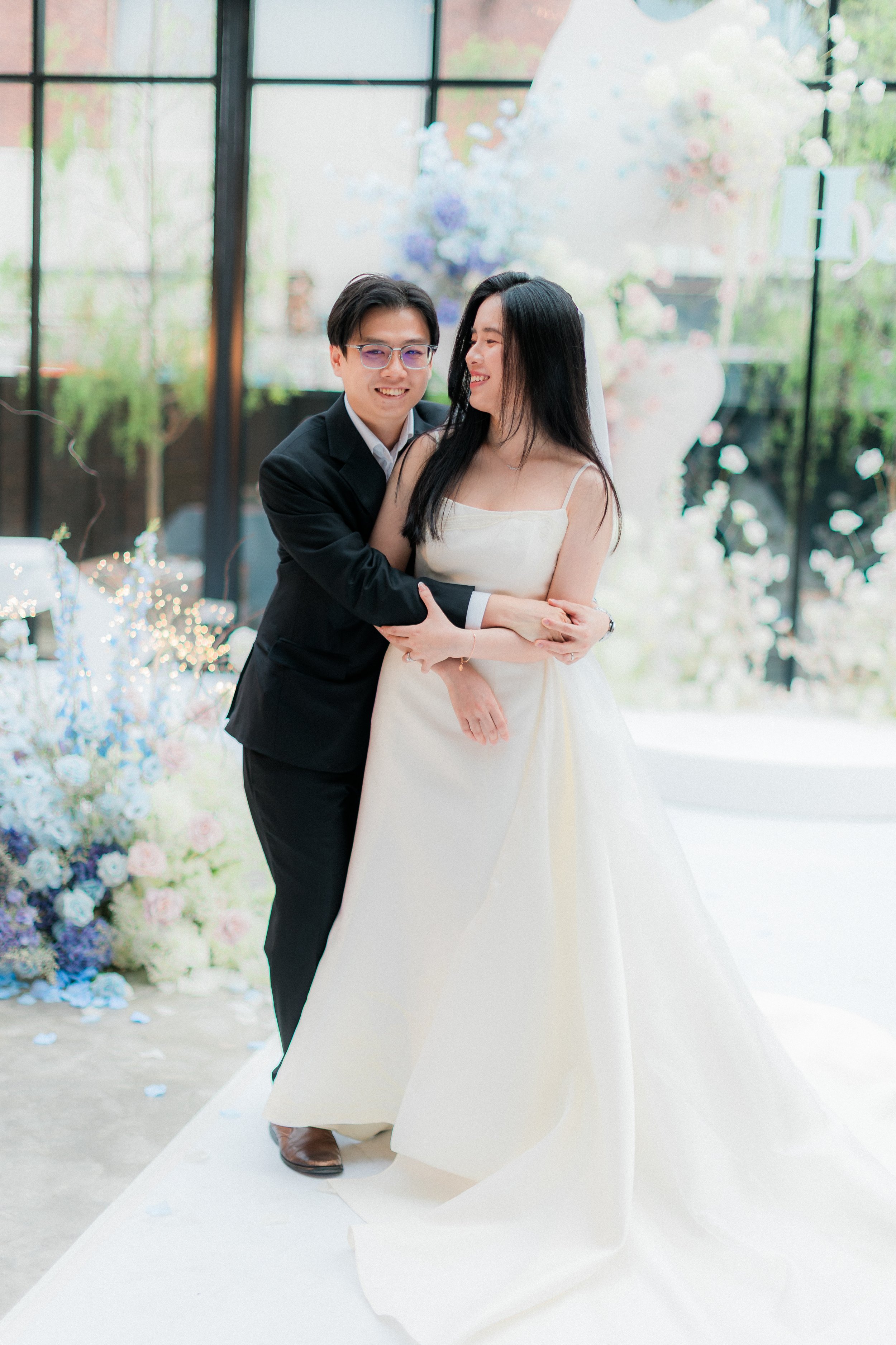 A bride and groom sharing a joyful moment at their wedding, standing indoors with large windows and floral decorations in the background.
