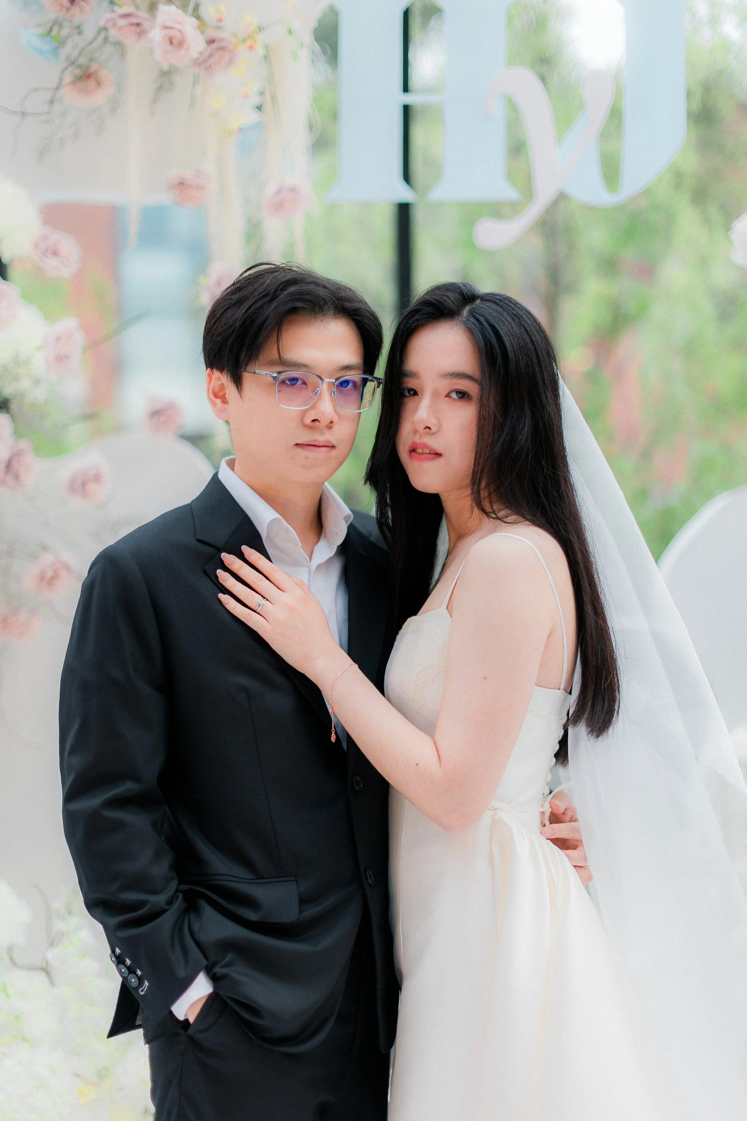 A couple dressed in wedding attire standing indoors with greenery visible outside the window behind them.