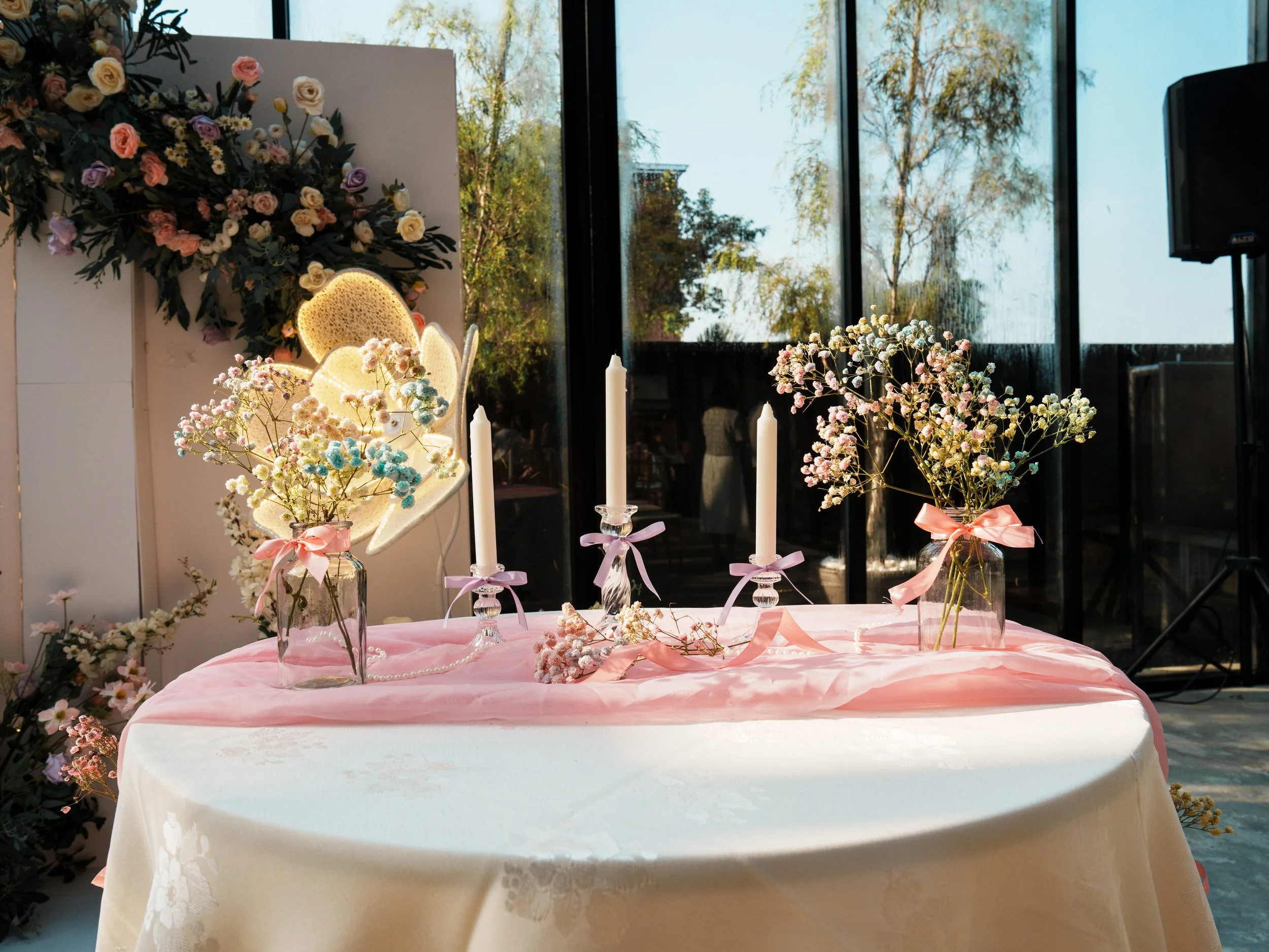 Decorative table set with pink and white flowers in glass jars with pink ribbons, white candles with pink ribbons, and a pink table runner, with a floral backdrop in a well-lit room with large windows.