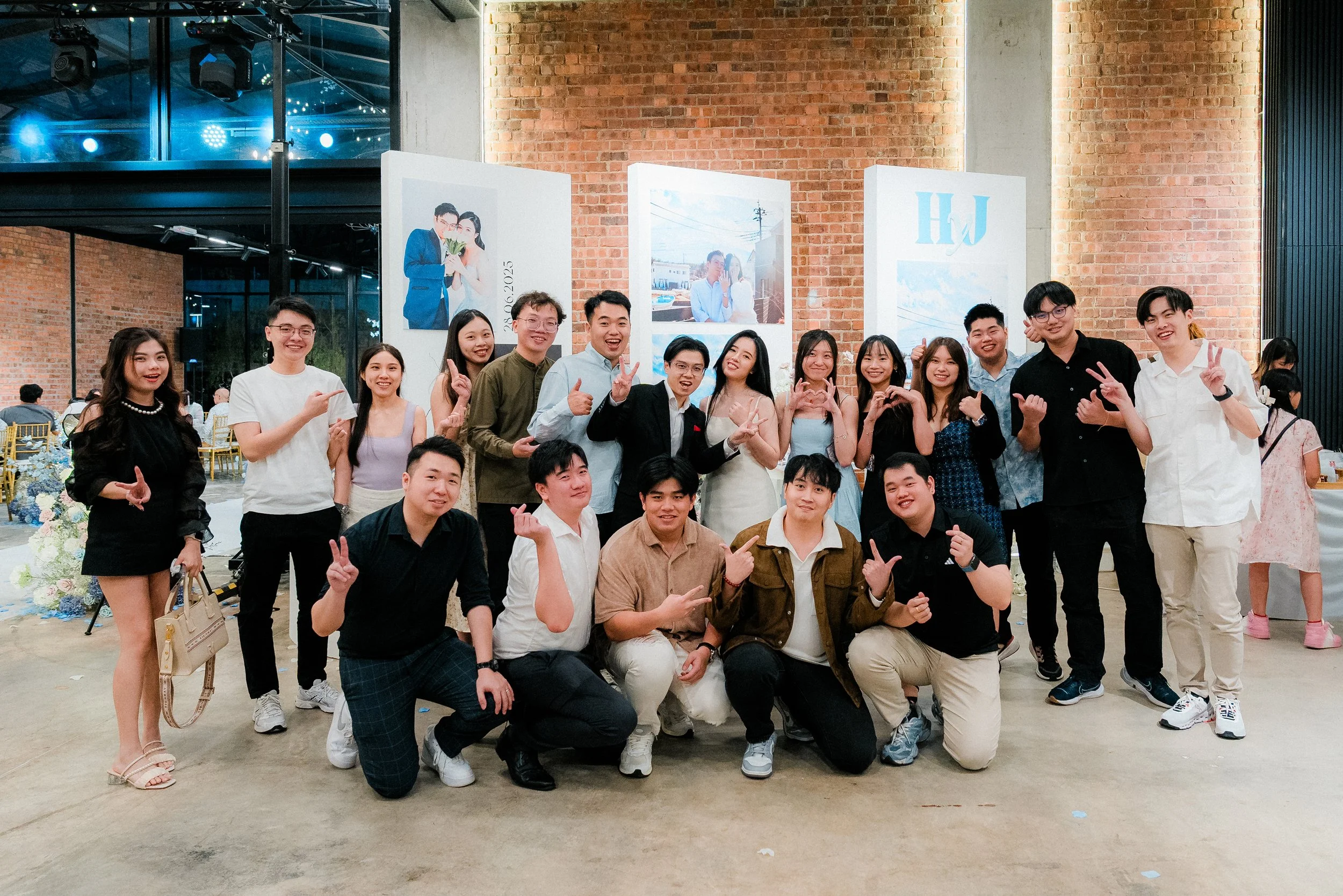 A large group of young adults posing and smiling for a photo at a celebration event inside a decorated venue with brick walls, tables, and large photo displays in the background.