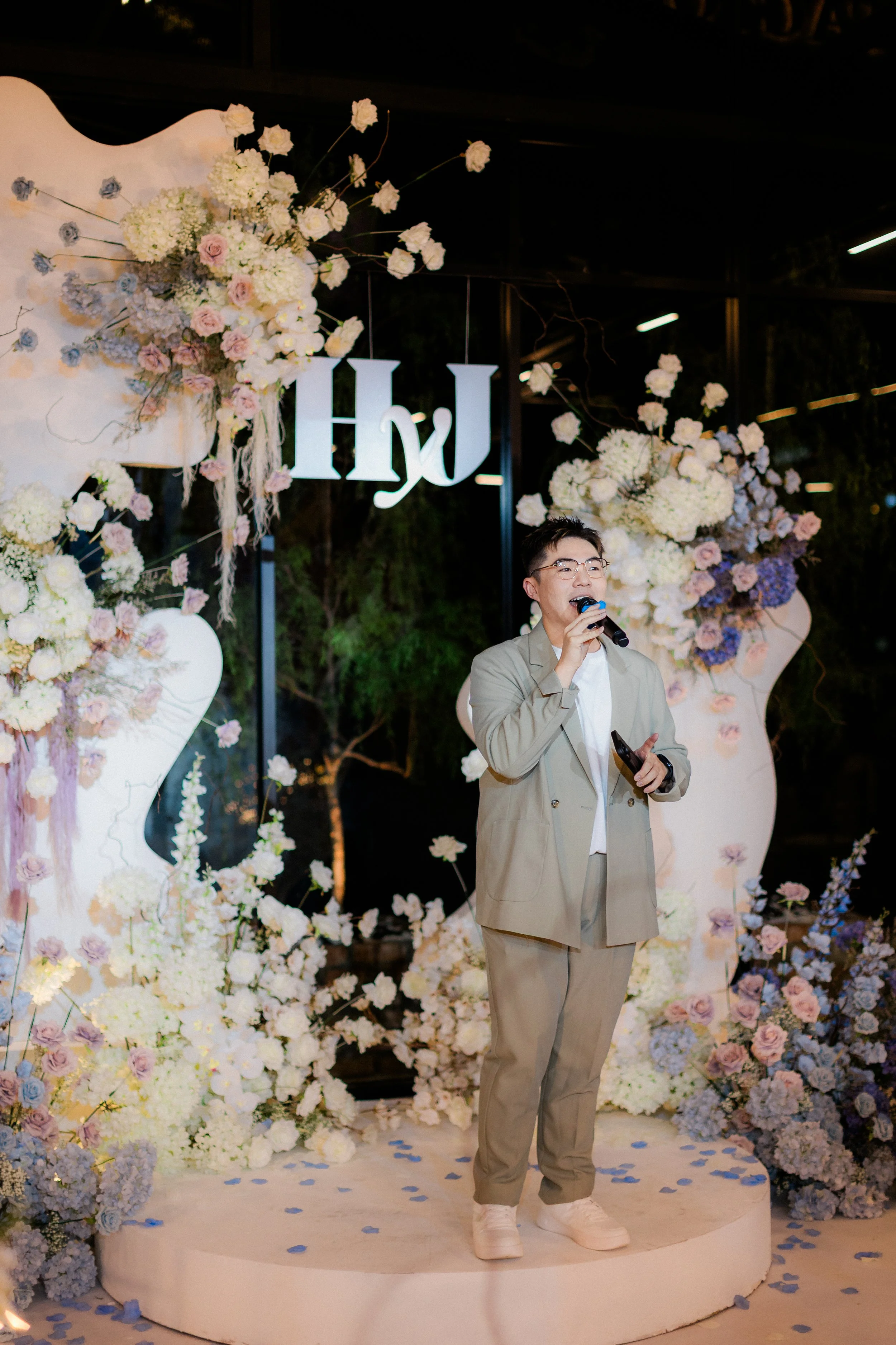 A person in a beige suit holding a microphone, standing on a decorated stage with white and pastel-colored flowers and the initials 'HW' hanging behind them.