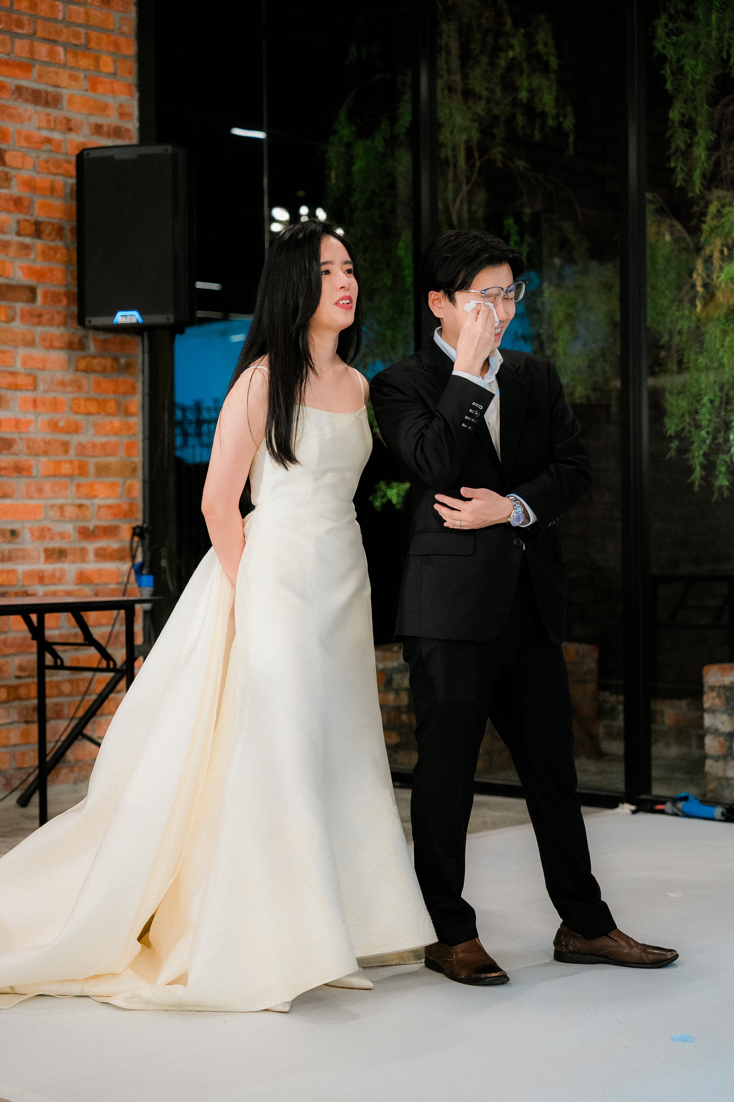 A bride in a white wedding dress and a groom in a black suit standing indoors near a brick wall and large glass windows, with the groom wiping tears from his eyes.