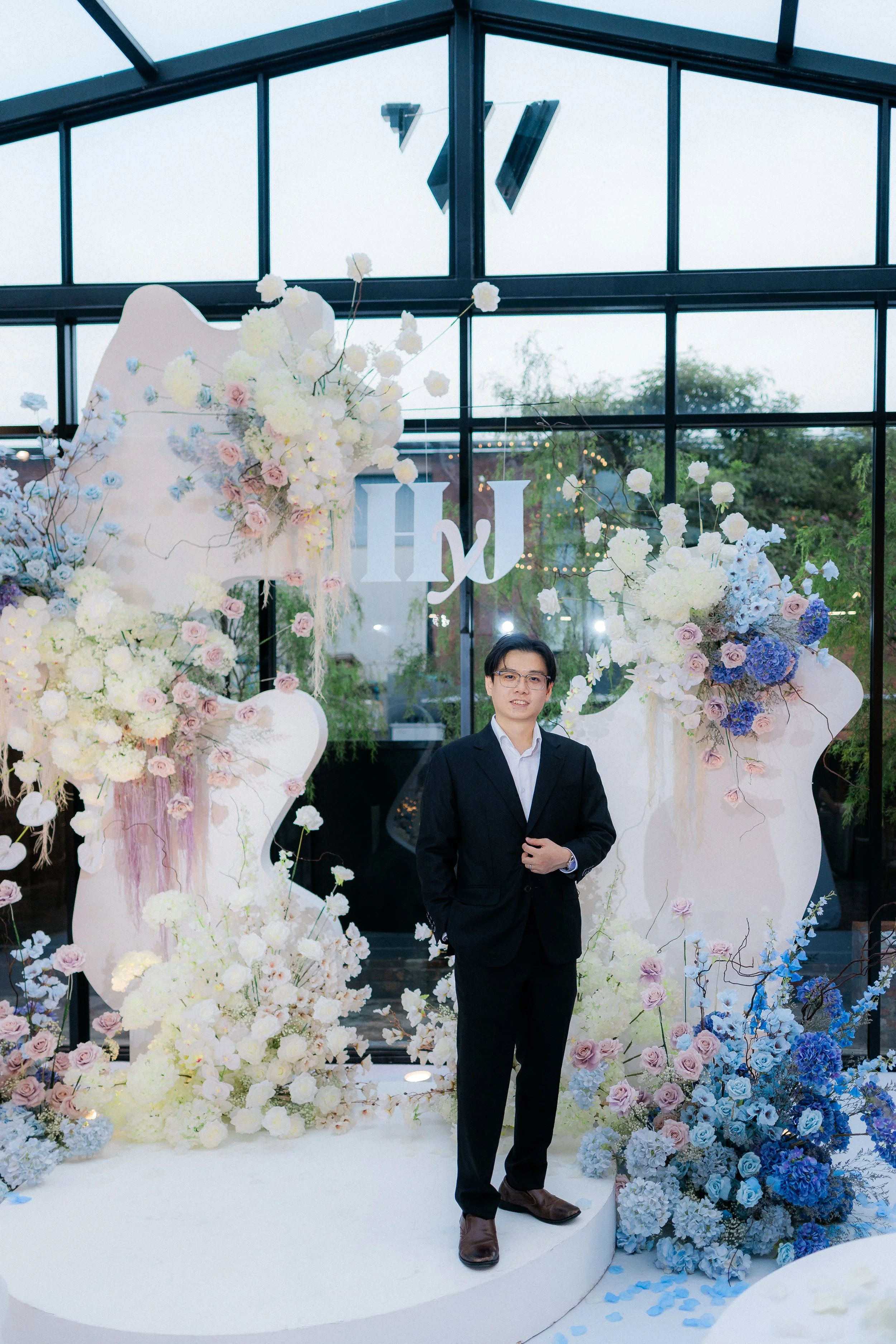 A man dressed in a black suit and glasses standing in front of a floral decoration with white, purple, and blue flowers, inside a glass building with trees visible outside.