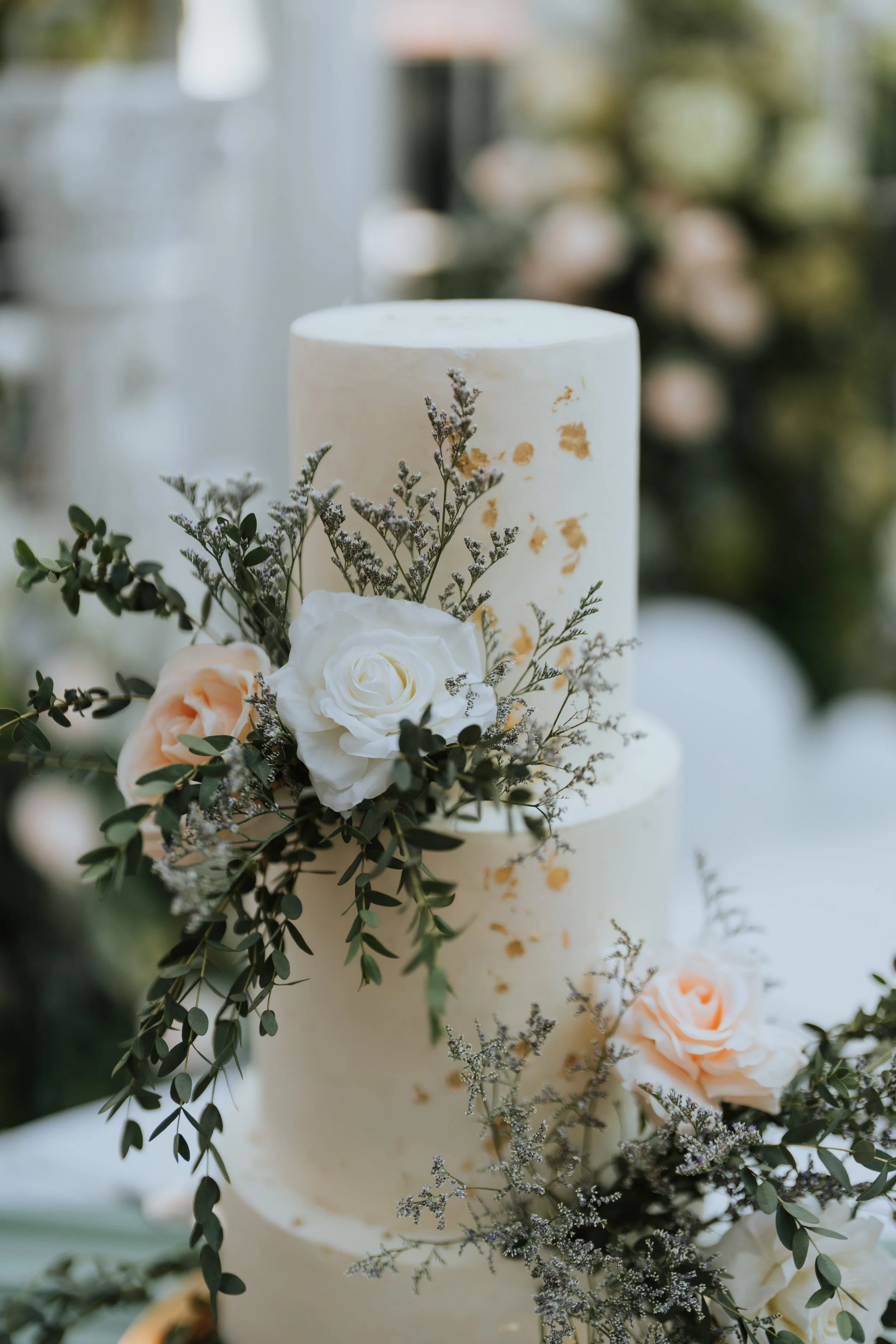 Elegant three-tier wedding cake decorated with white and peach roses, greenery, and gold accents.
