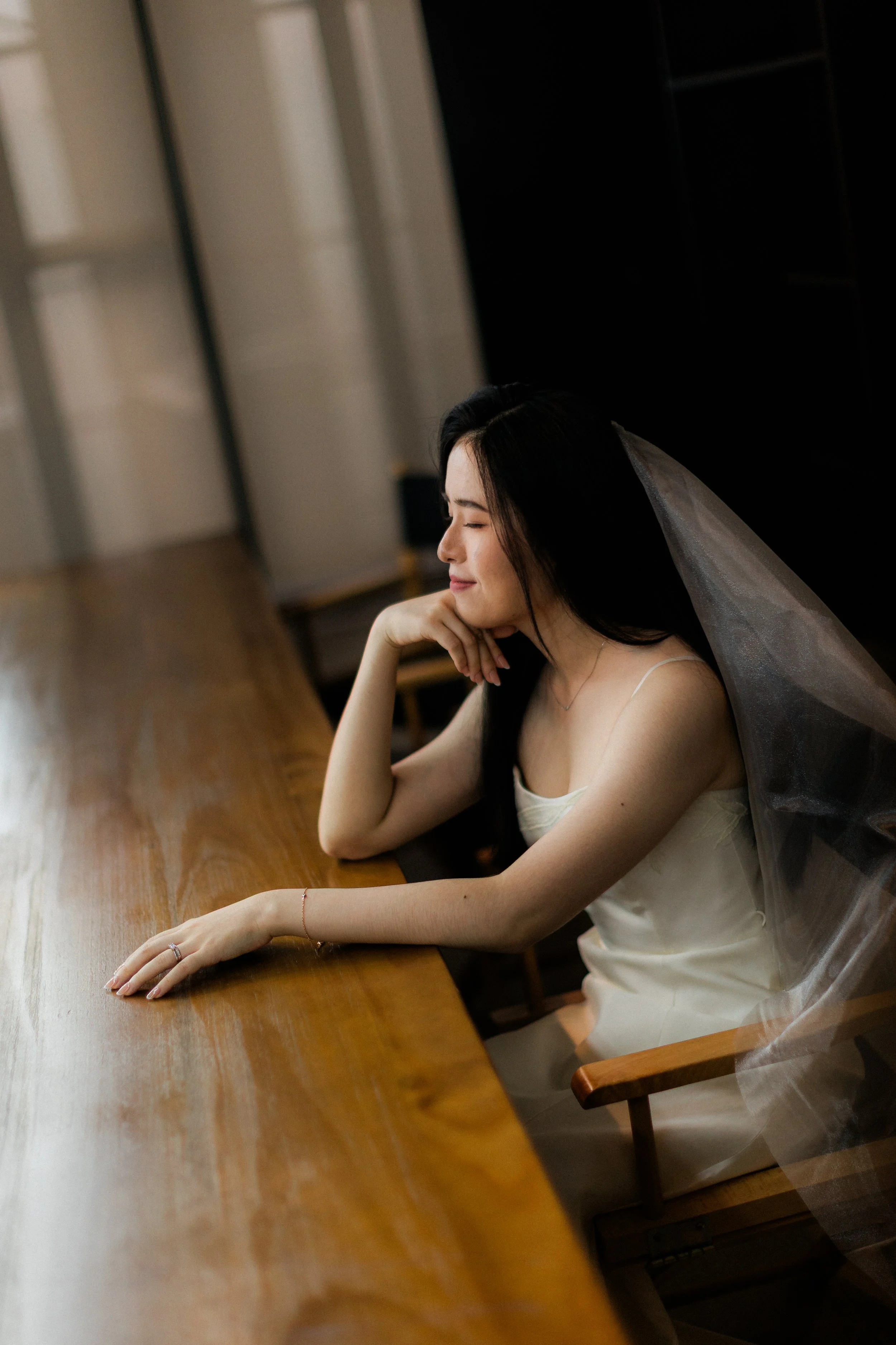 A young woman with long black hair wearing a white dress and a wedding veil, sitting at a wooden table with her eyes closed and a smile, resting her chin on her hand.