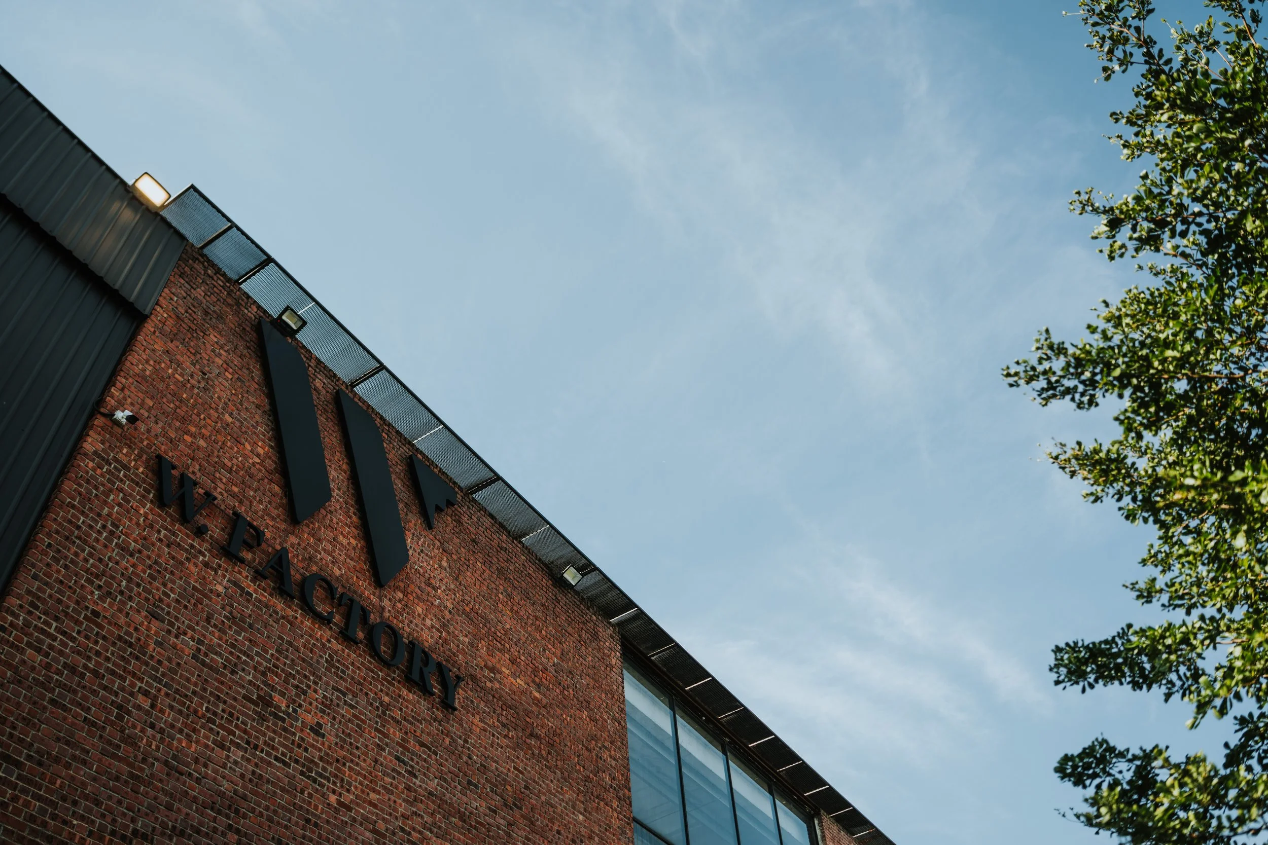 A brick building with the words "W. Pactory" on it, large black logo, and a clear blue sky with some clouds, partially visible tree on the right.