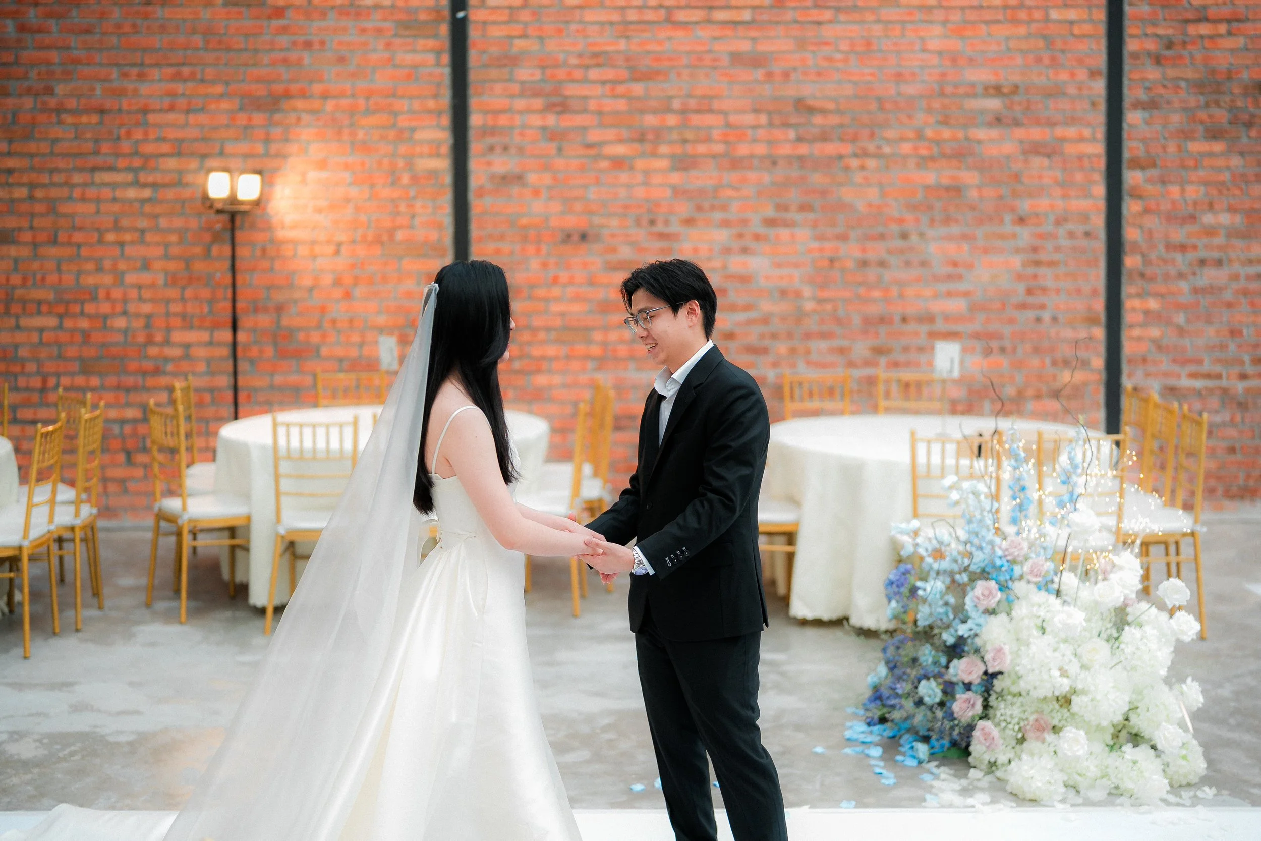 A bride and groom holding hands during a wedding ceremony in a decorated venue with a brick wall background and floral arrangements.