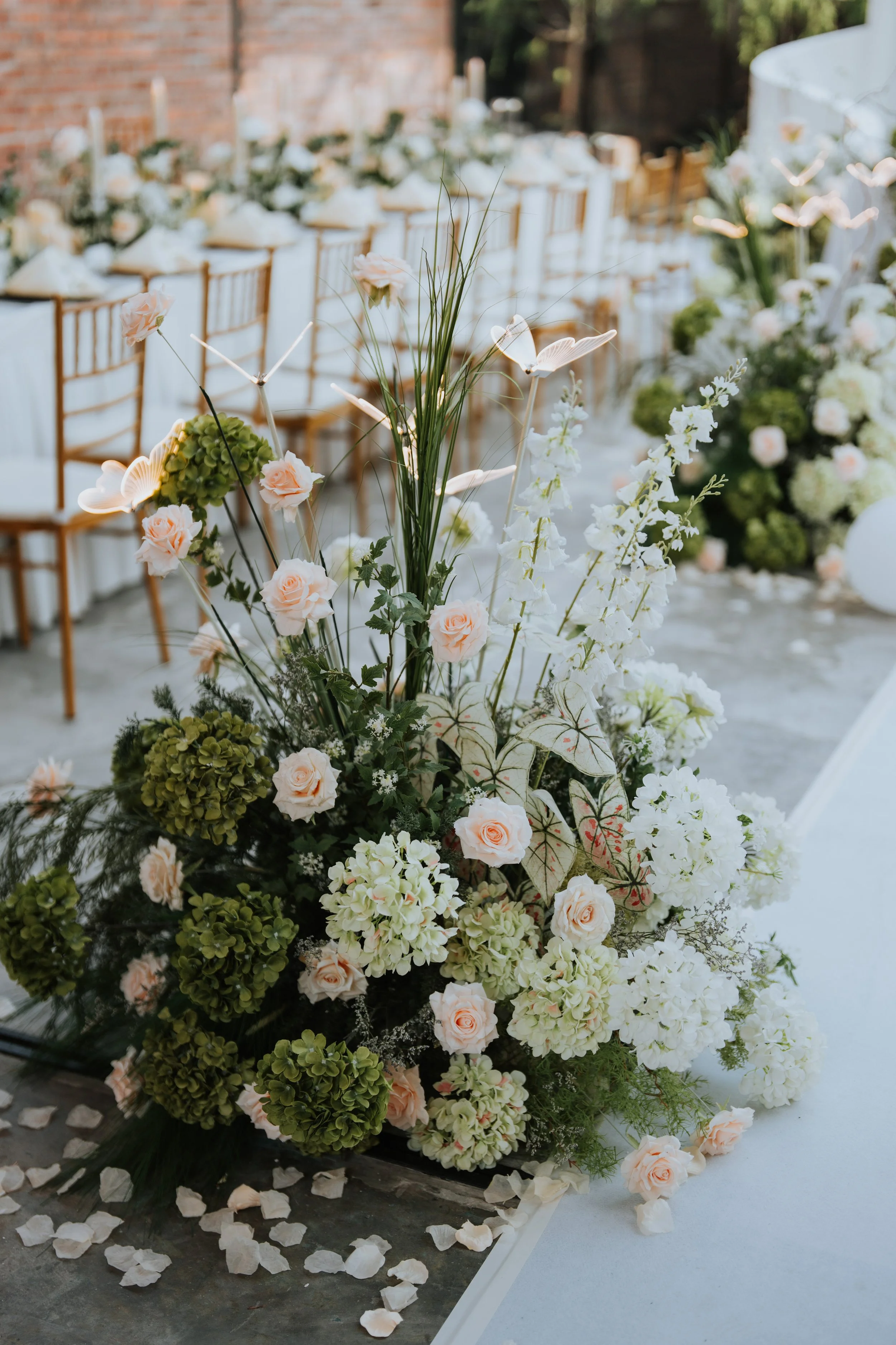 Elegant floral centerpiece with white and peach roses, green hydrangeas, white orchids, and greenery on a table with scattered petals, set for a wedding reception.