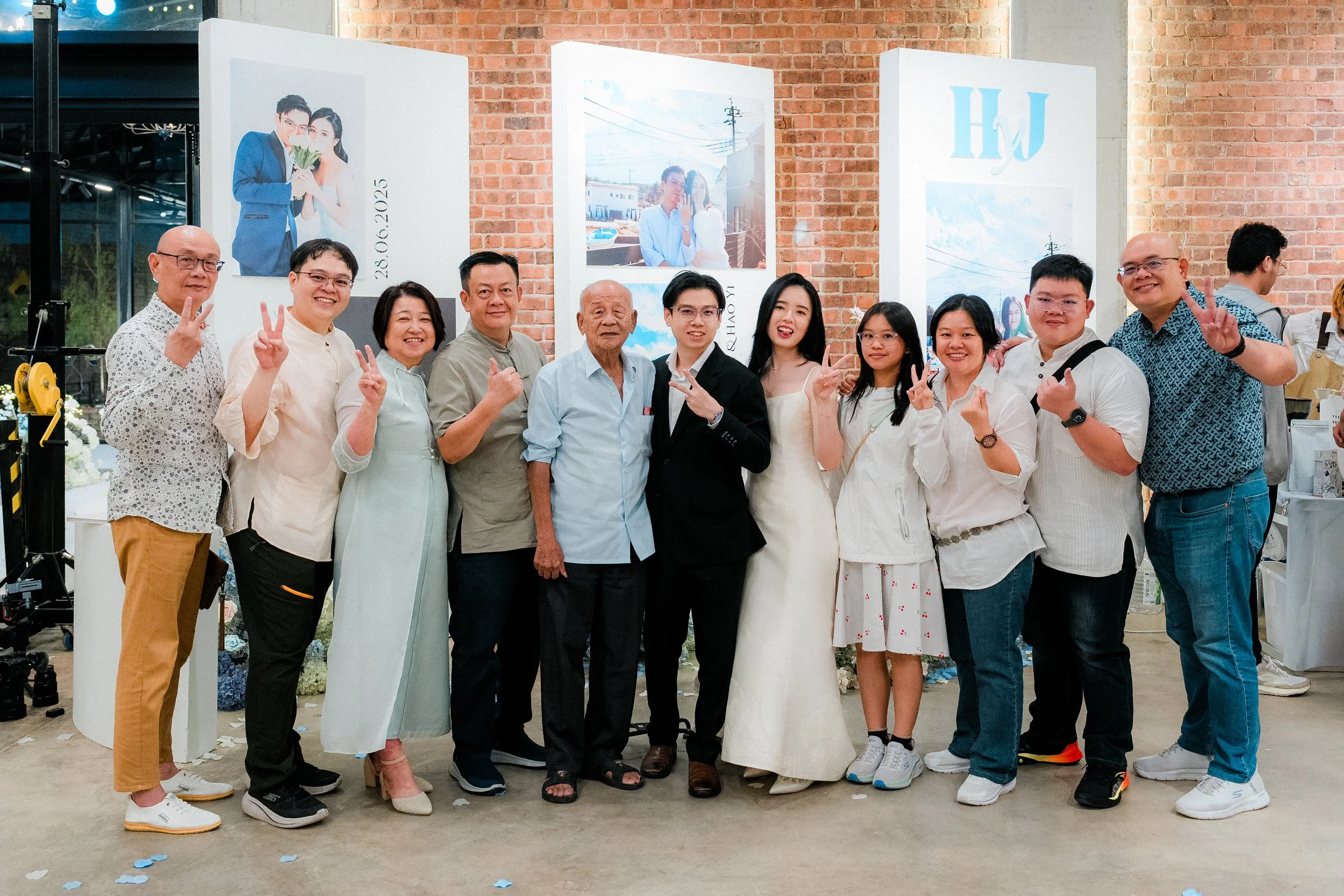Group of people posing for a photo at a celebration or wedding event, with some making peace signs. There are large photo displays in the background with images of a couple and a brick wall.