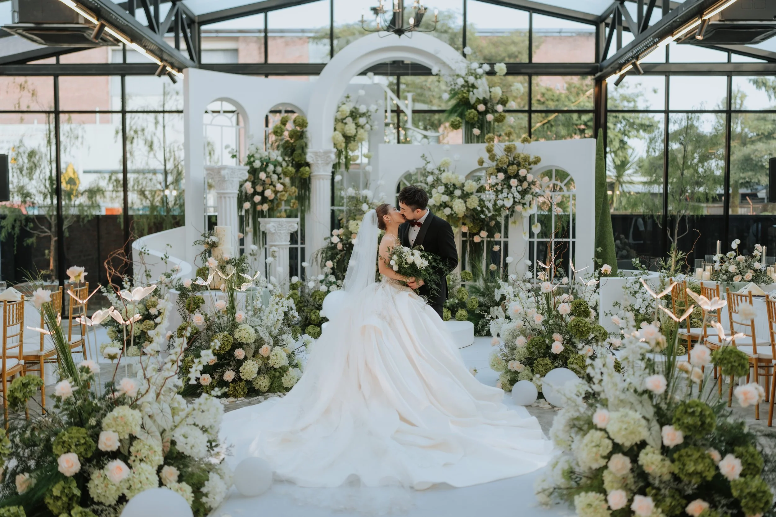 A bride and groom share a kiss at their wedding ceremony inside a glass-walled venue decorated with white flowers, arches, and greenery.