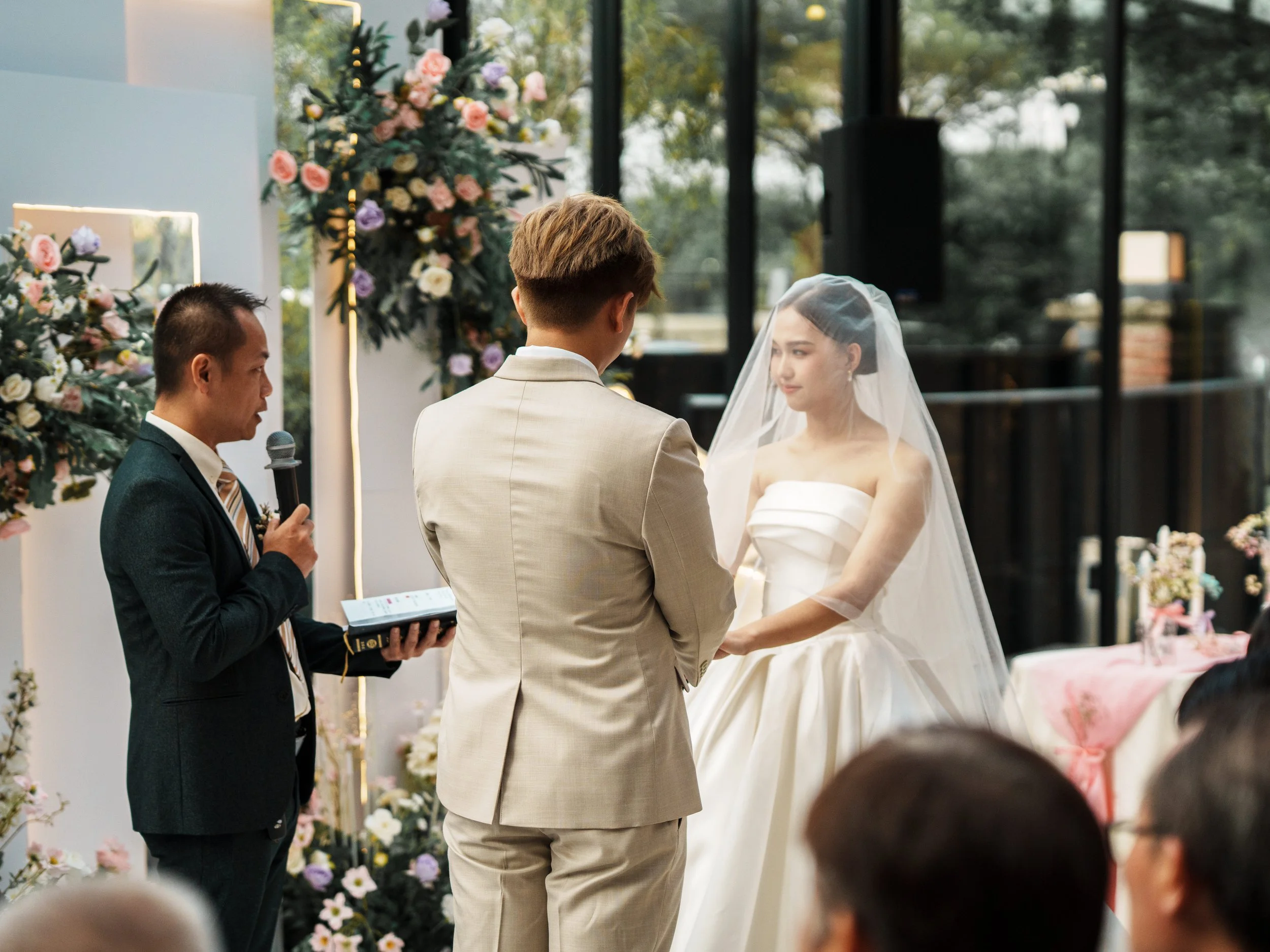 A wedding ceremony taking place indoors with a bride in a white strapless wedding gown and veil, standing opposite a groom in a beige suit, holding hands. An officiant in a dark suit is reading from a book into a microphone, with floral decorations i