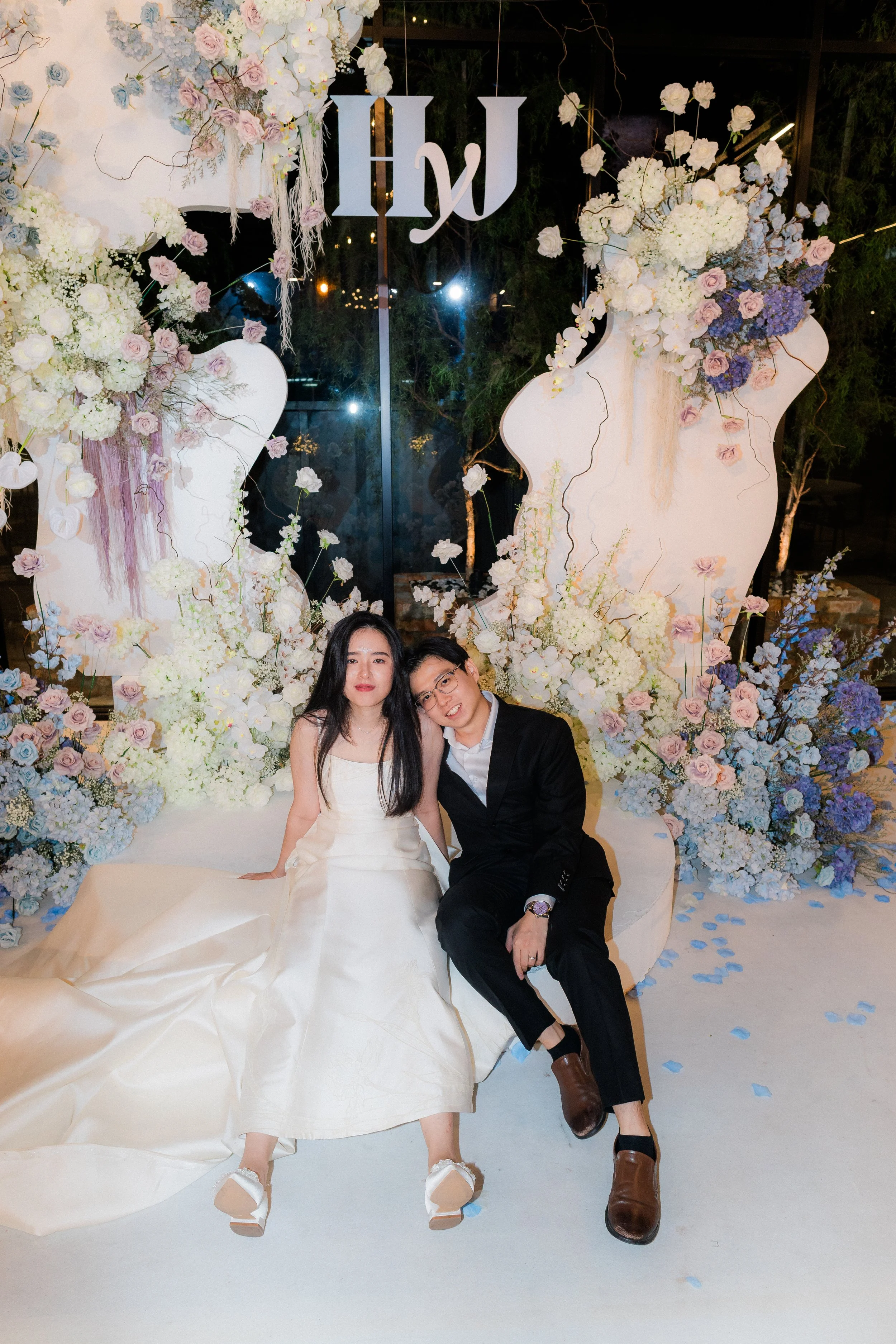 A couple in formal attire sitting on a white sofa with a floral backdrop at a wedding or event. The woman is in a white gown, and the man is in a black suit. There are floral arrangements and the word 'Hiyu' hanging overhead.
