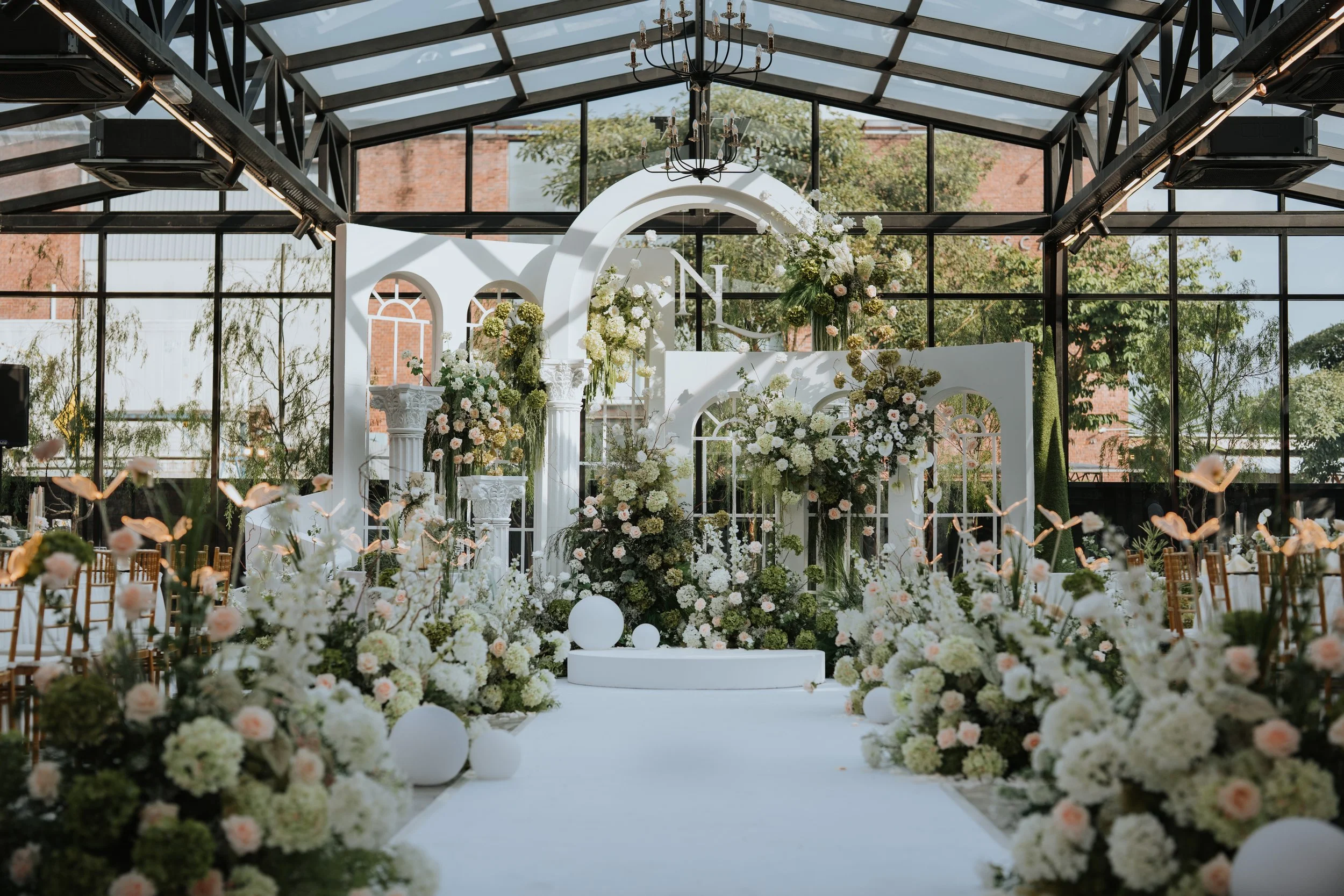 Elegant indoor wedding altar decorated with white and pastel-colored flowers, white pillars, and circular architectural features, inside a glass-roofed venue with outdoor views.