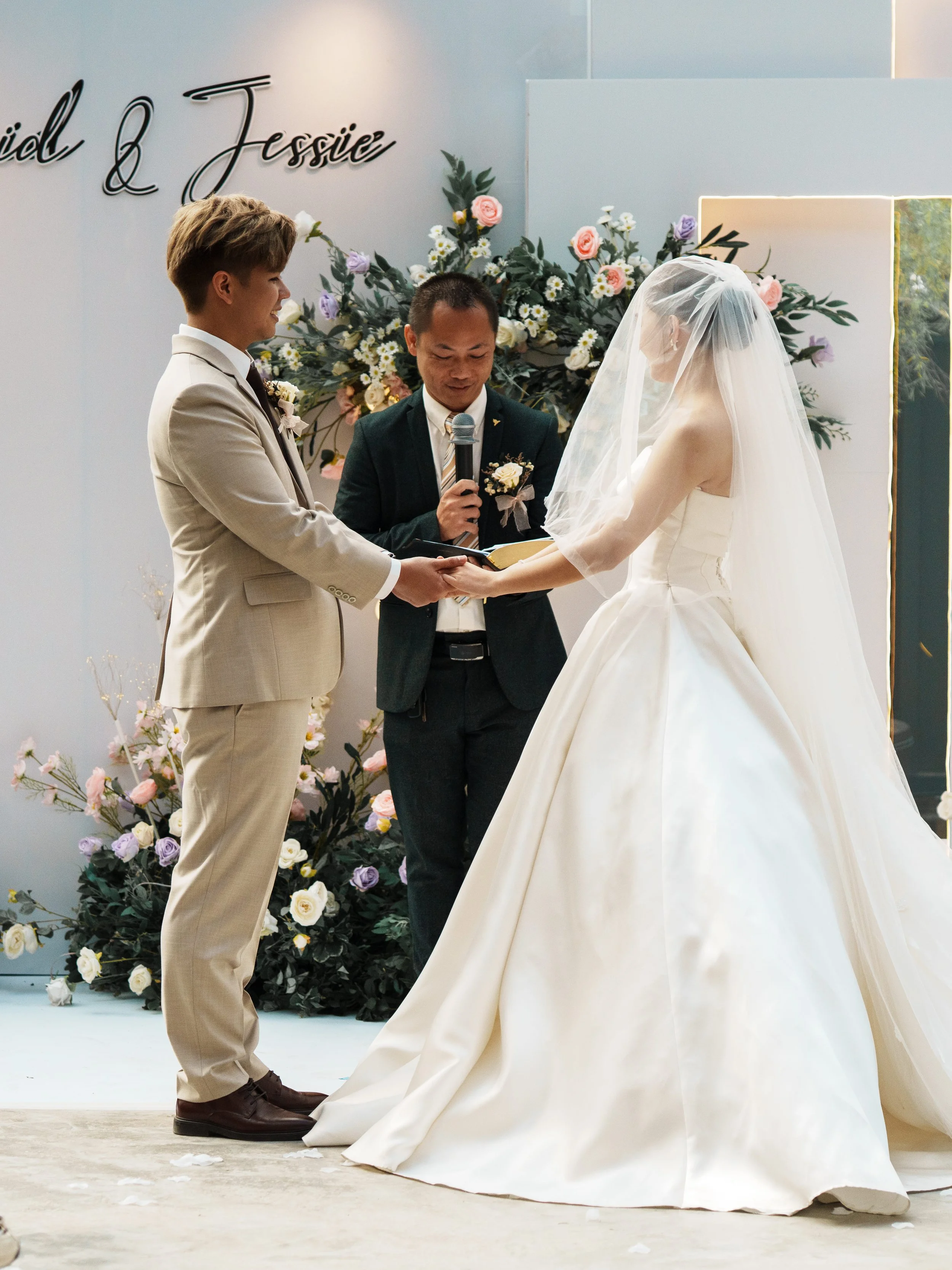 A bride and groom holding hands during their wedding ceremony with an officiant reading vows, in front of floral arrangements with pink, purple, and white flowers.