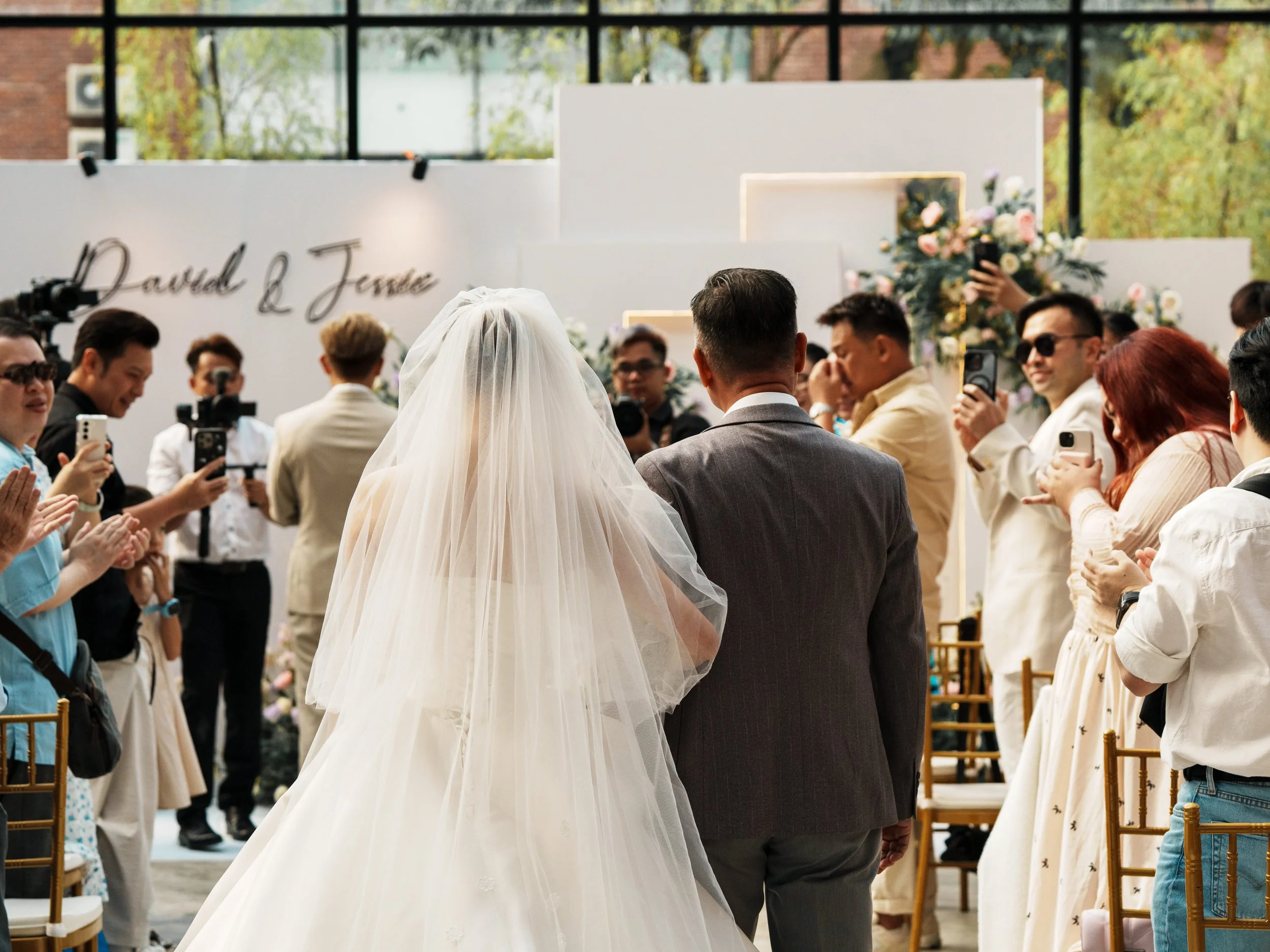 Bride in a white wedding gown and veil walking down the aisle with a man in a gray suit, surrounded by seated guests taking photos and clapping at a wedding ceremony