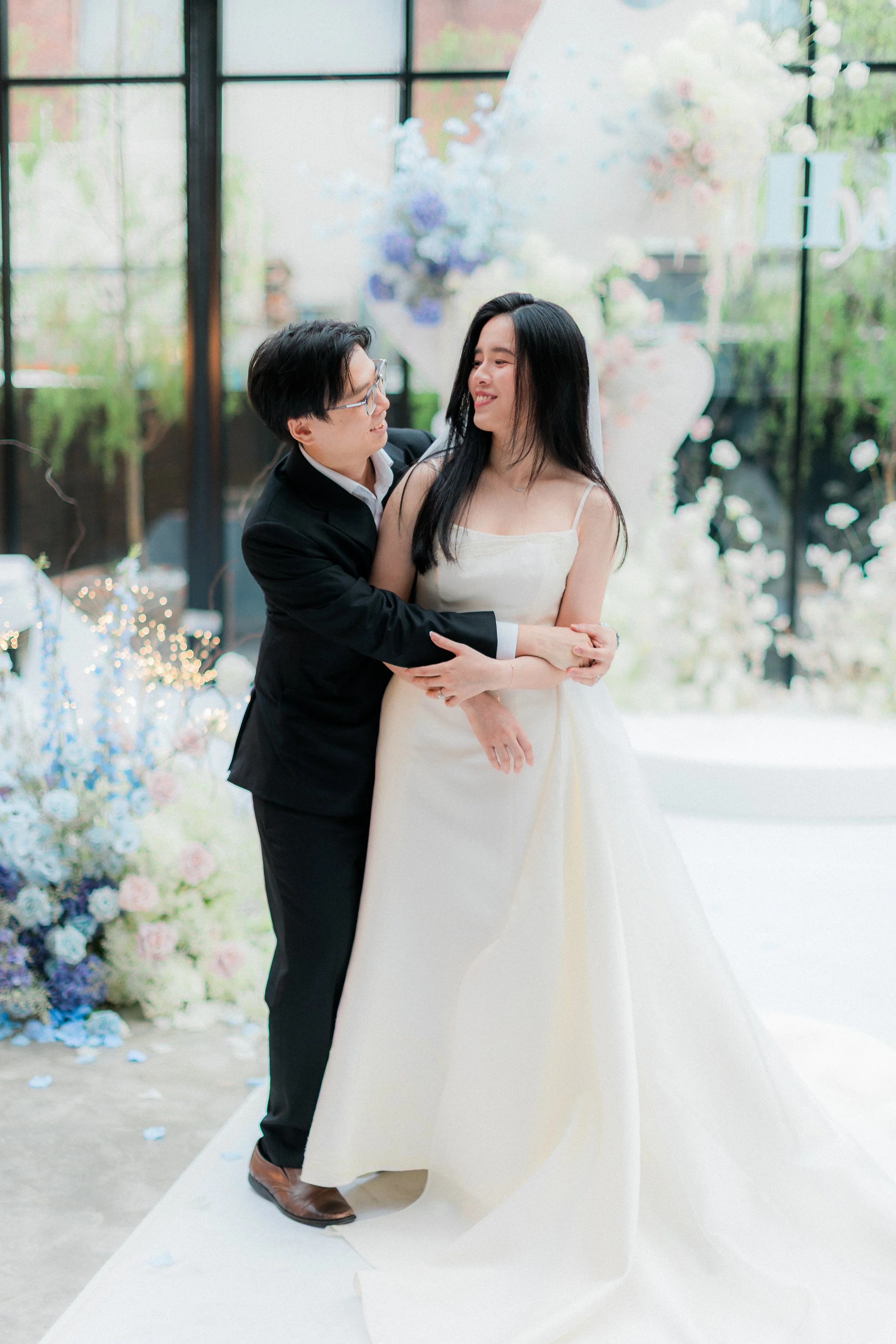 A couple sharing a tender moment at a wedding, with floral decorations and large windows in the background.