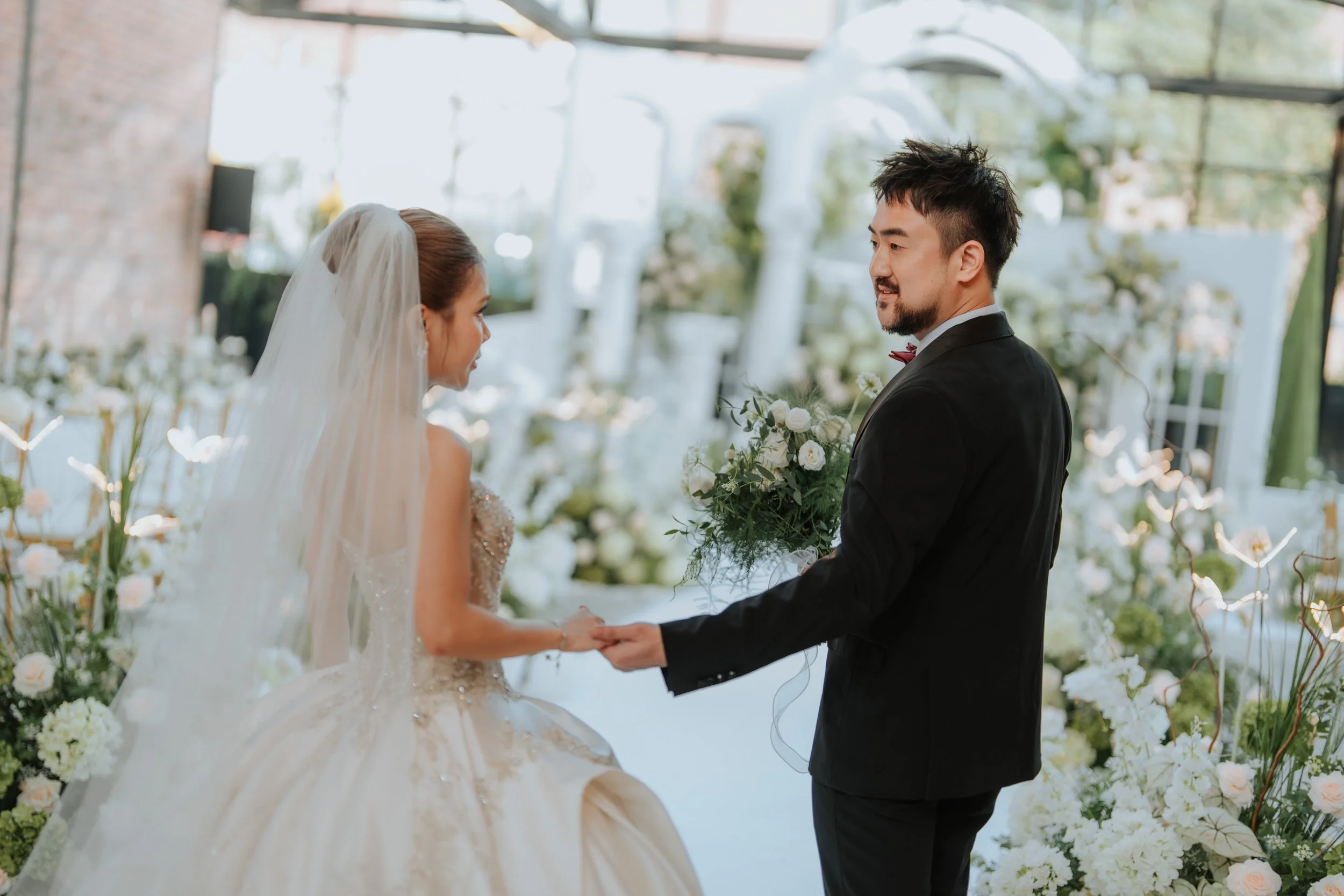 Bride and groom holding hands during wedding ceremony with floral decorations in the background.