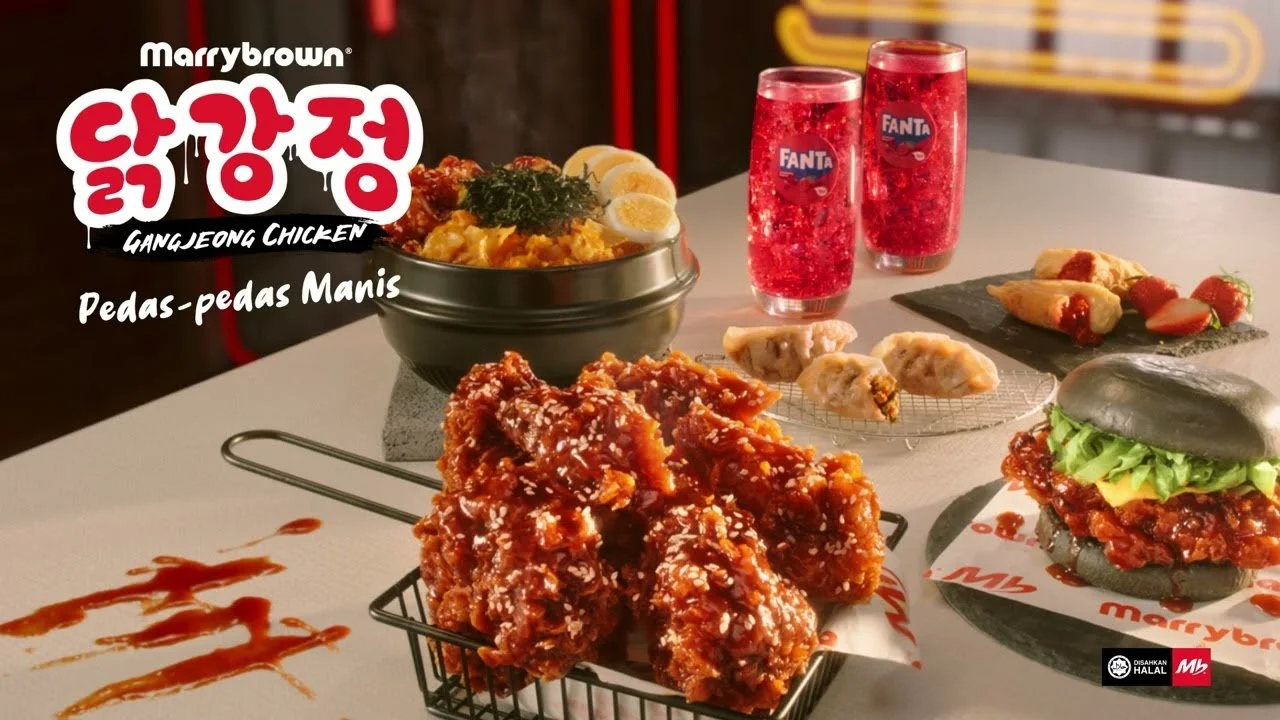 Table with Korean fried chicken, two glasses of Fanta, a bowl of rice with toppings, a small plate of appetizers, and a burger with black bun and lettuce.