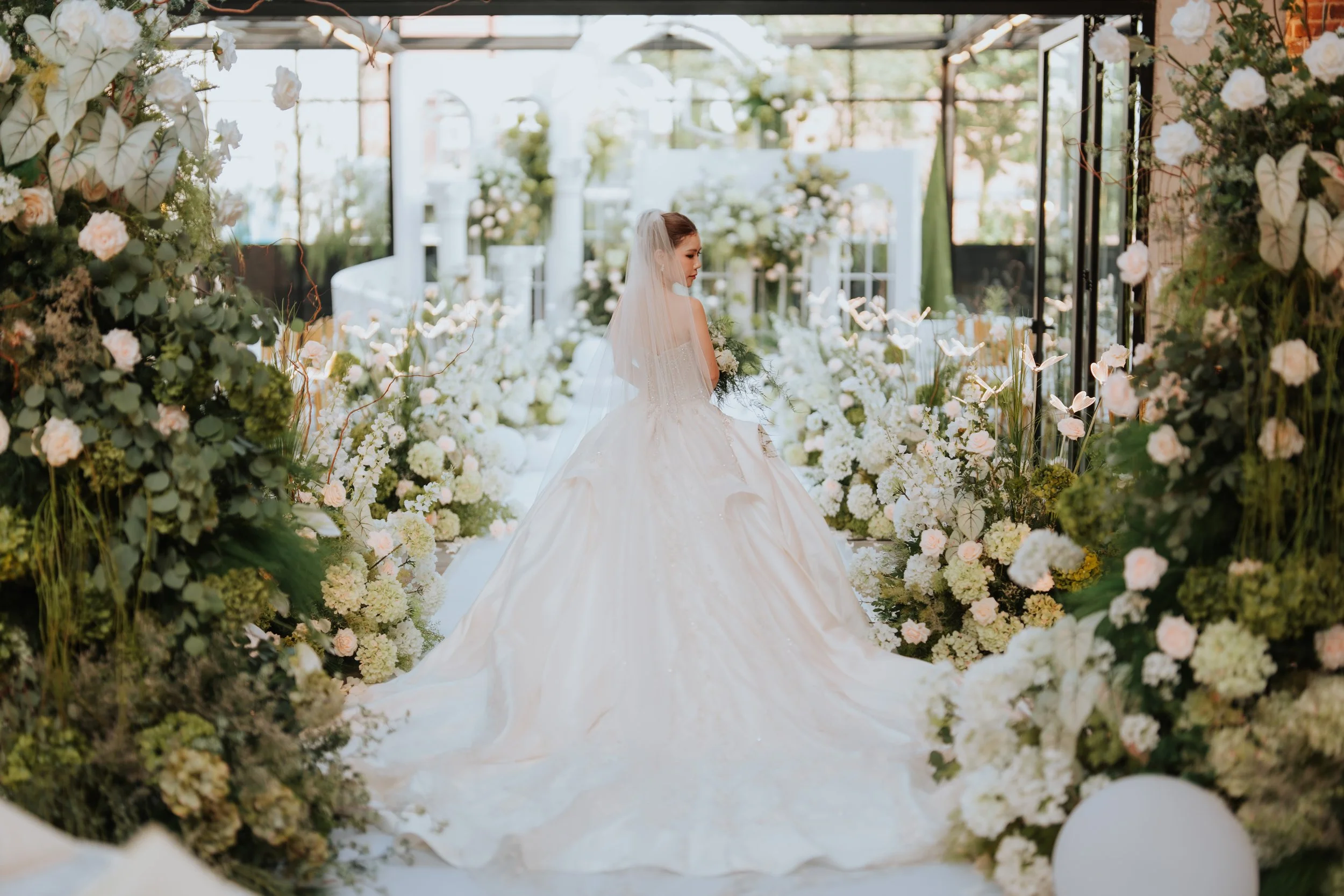 A bride in a white wedding gown with a long train, standing in an aisle decorated with white flowers and greenery at a wedding venue.
