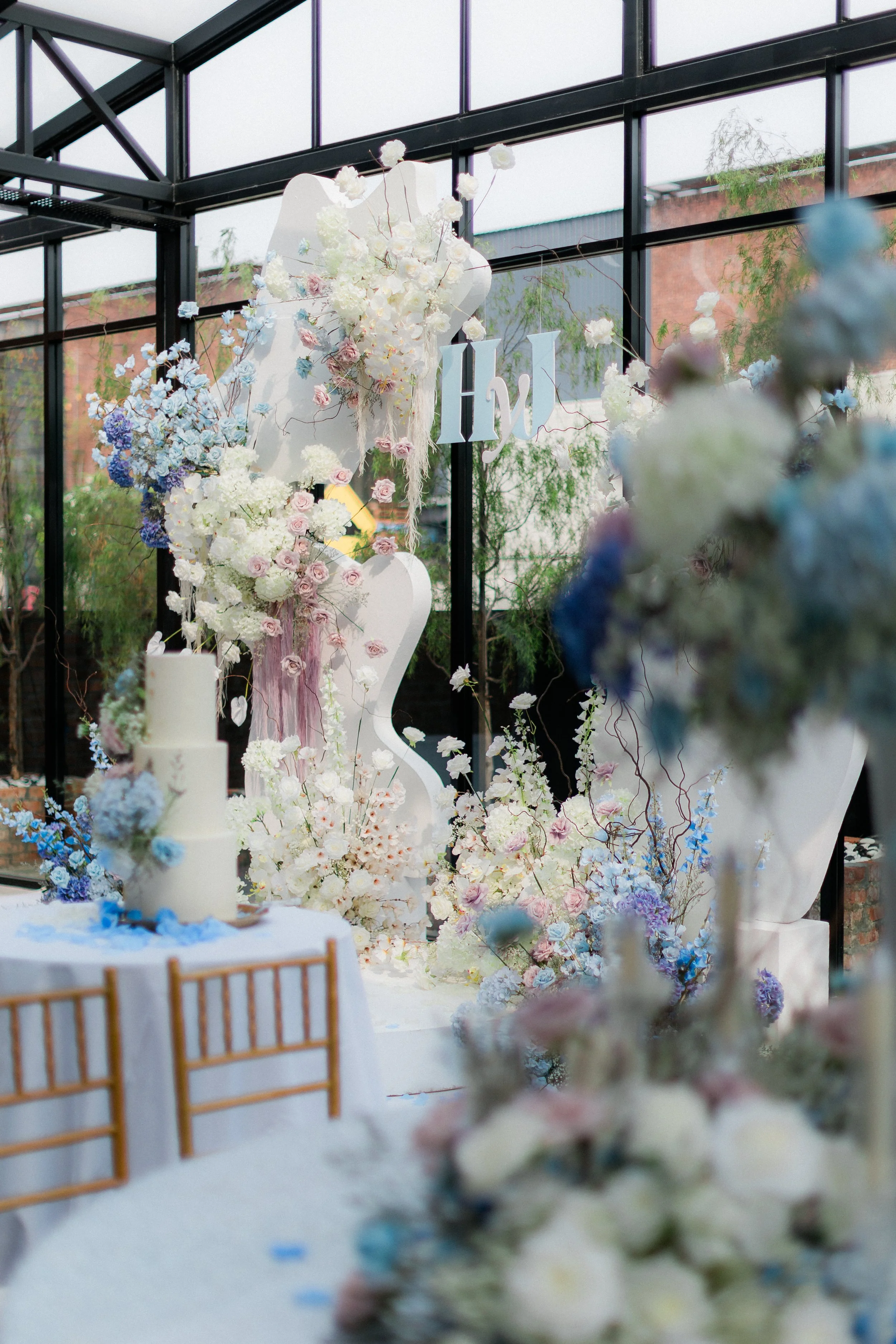 Elegant indoor wedding reception with floral arrangements, a wedding cake, and decorative letters 'h' and 'l' in a glass conservatory.