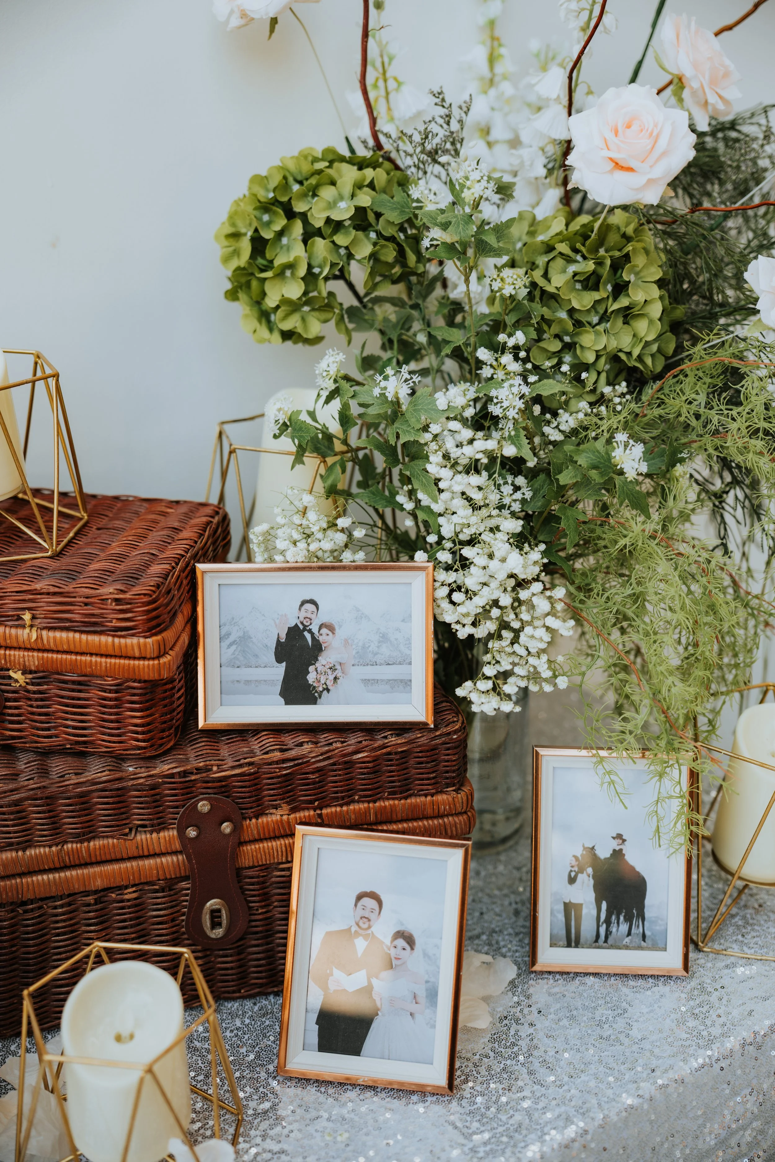 Decorative display with framed wedding photos, white candles in gold geometric holders, a woven wicker box, and a backdrop of white and green floral arrangements including roses, hydrangeas, and greenery on a sequin tablecloth.