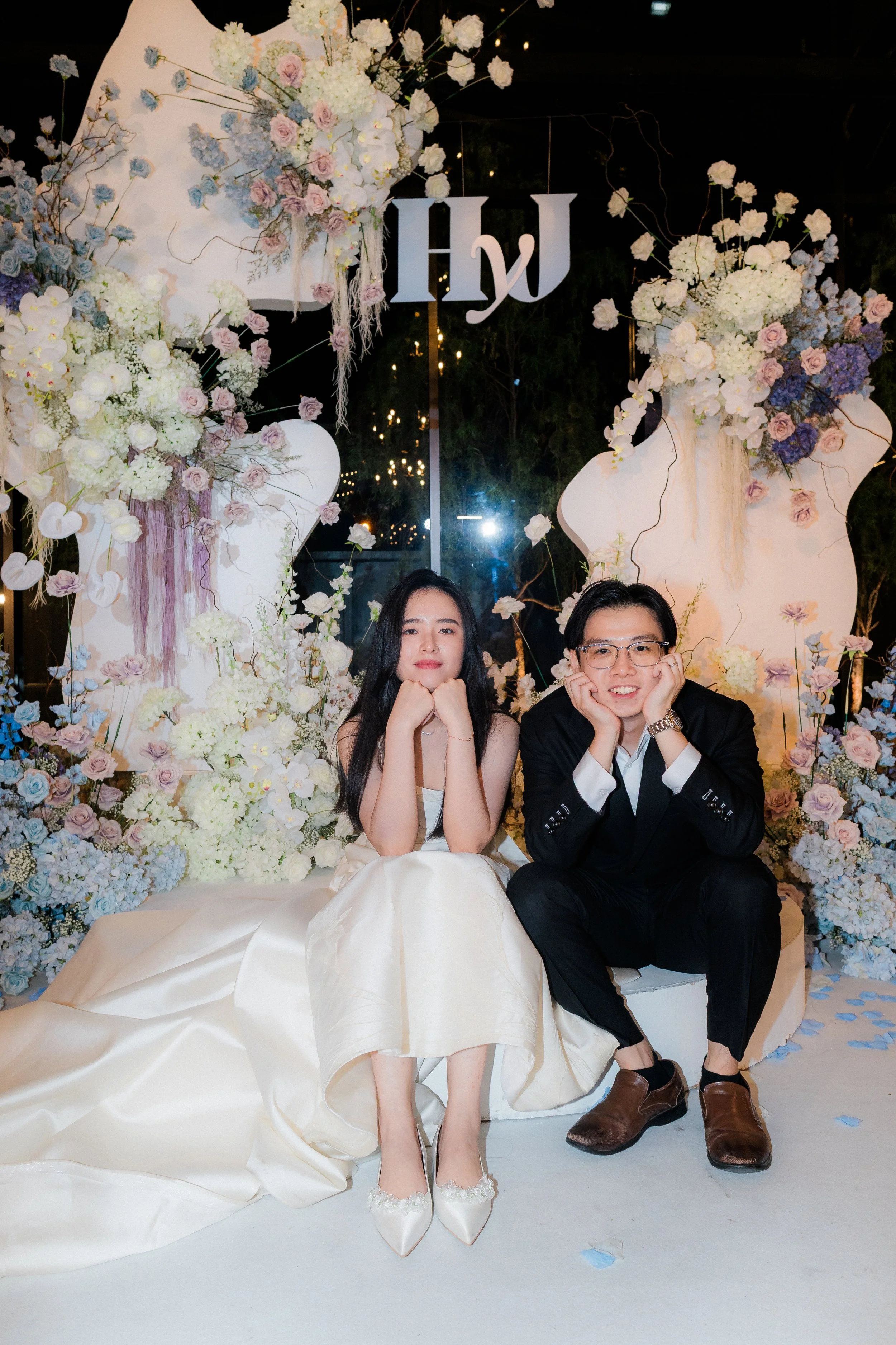 A couple sitting on a floral decorated backdrop, likely at a wedding or engagement event, with the woman wearing a cream-colored dress and the man in a black suit, both resting their chins on their hands and smiling.