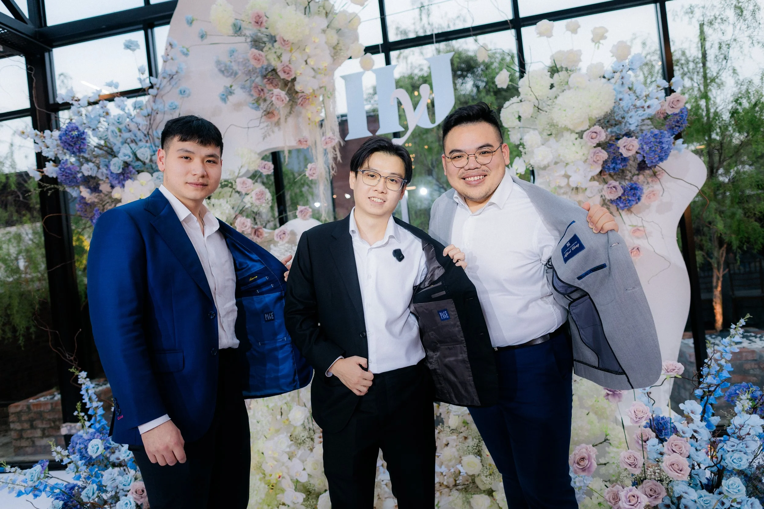 Three men in formal attire, smiling, showing off their jackets, standing in front of a floral backdrop with white and pink flowers, and a large letter 'H' and 'J' sign. The setting appears to be a celebration or event.