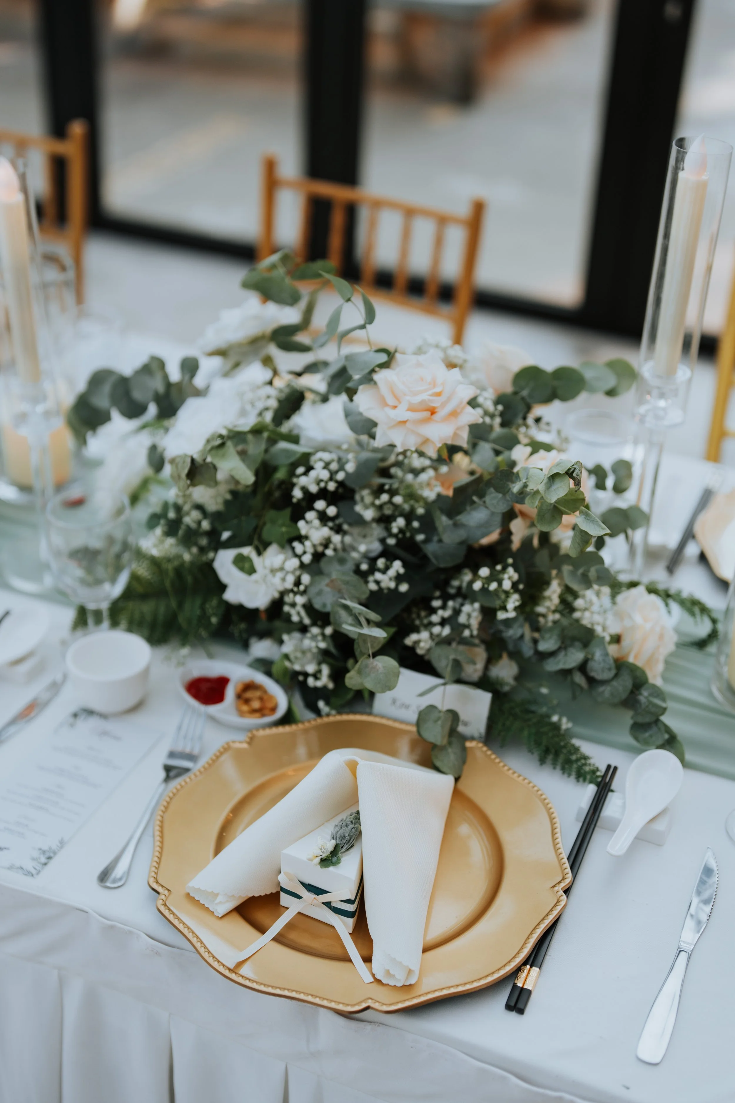 Elegant wedding table setting with a large floral centerpiece, gold charger plate, white napkin, small favor box, and candles.