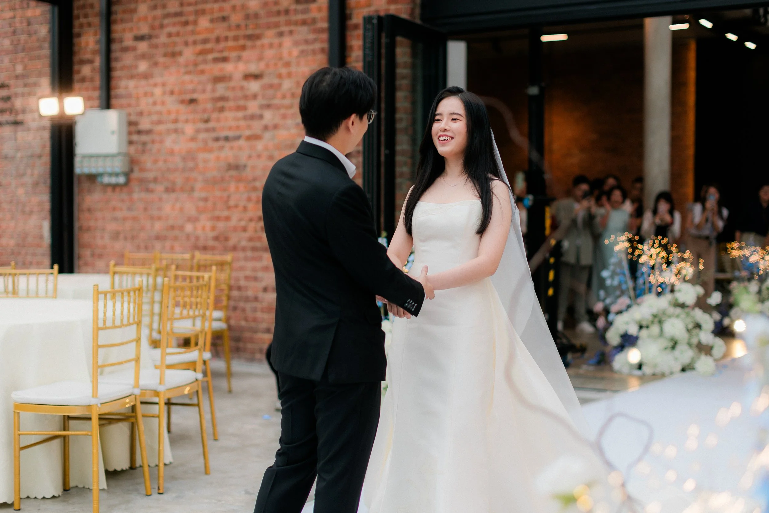 A bride and groom holding hands and looking at each other during their wedding ceremony in an industrial-style venue with brick walls and guests in the background.