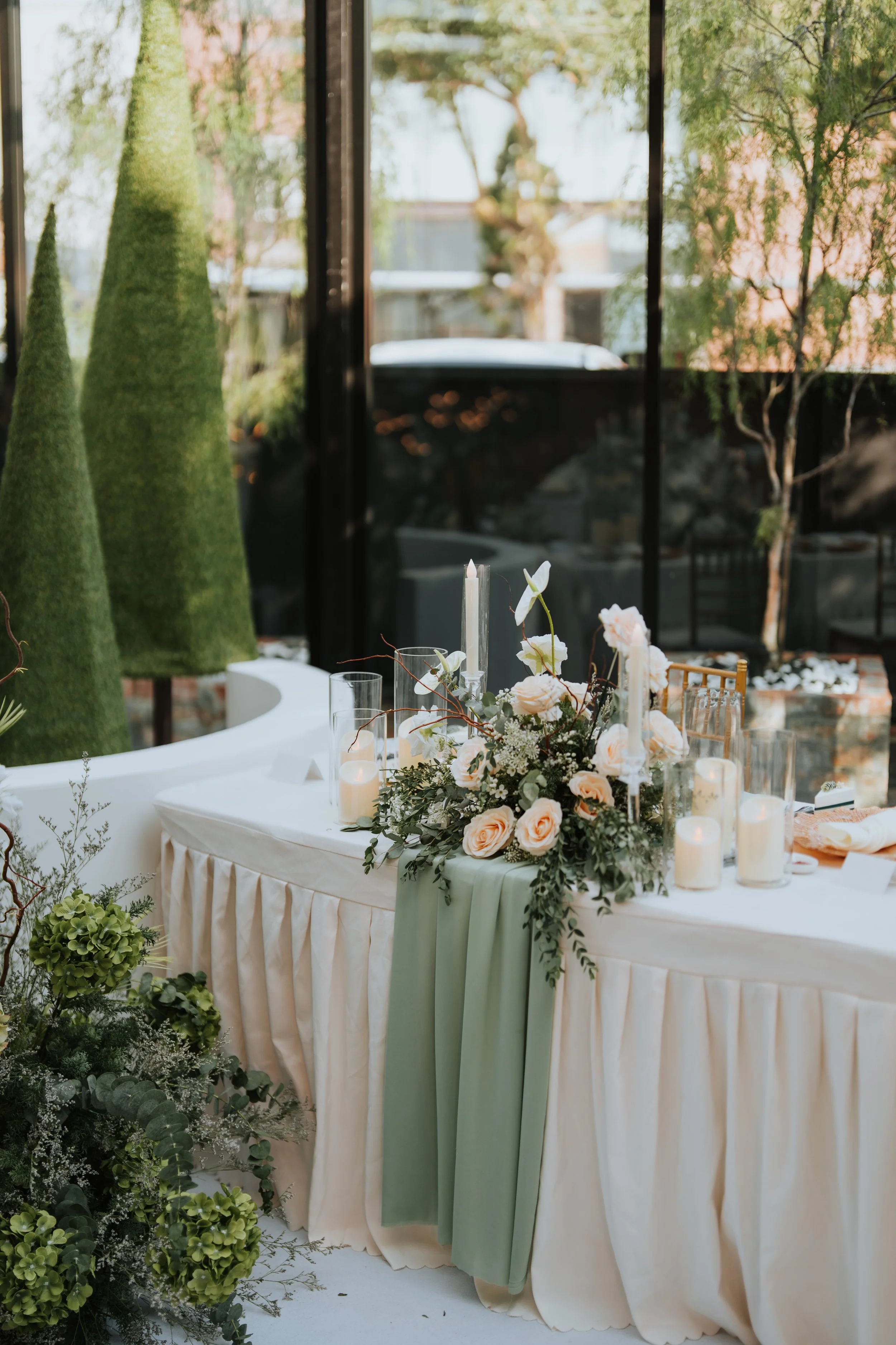 Elegant table decorated with white roses, candles, and greenery in a bright indoor space with large windows and outdoor greenery visible outside.