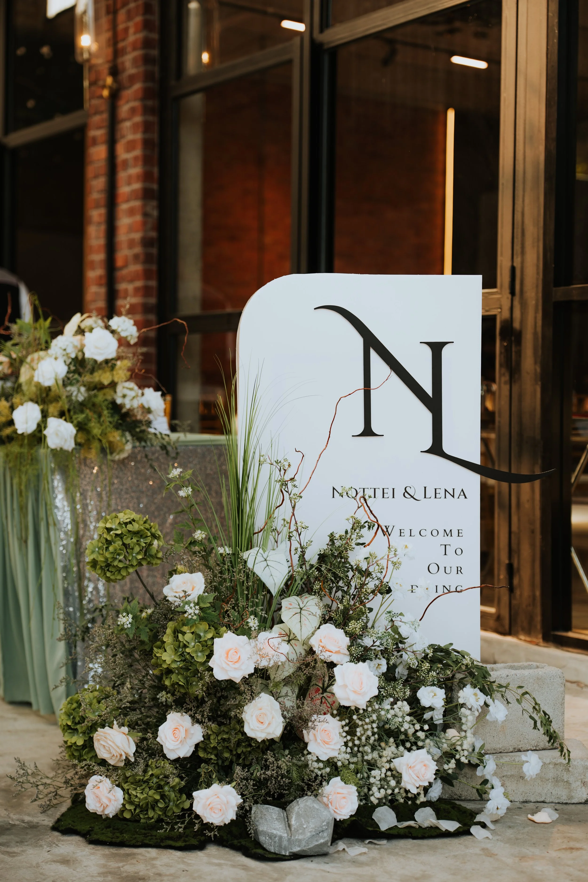 A white wedding sign with black lettering, decorated with white roses, green hydrangeas, and various greenery, placed in front of a rustic brick and metal building.