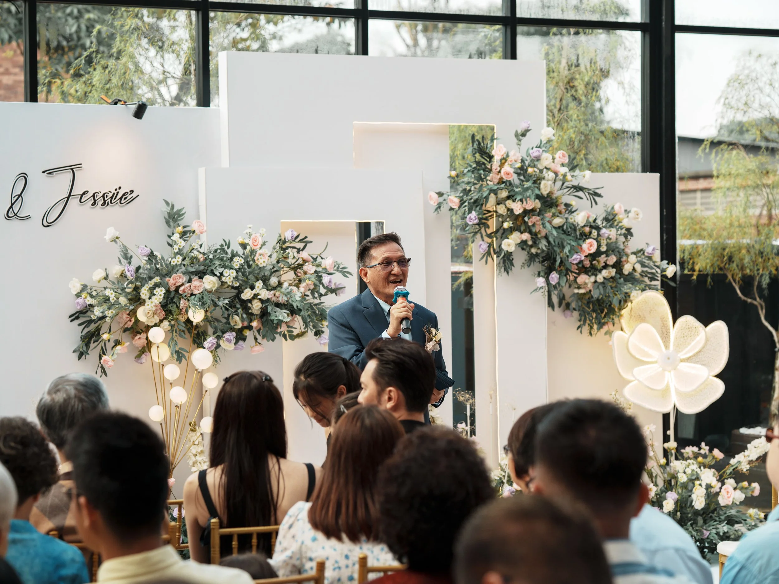 Man in blue suit speaking into microphone at wedding reception, surrounded by seated guests, with floral decorations and white backdrop.