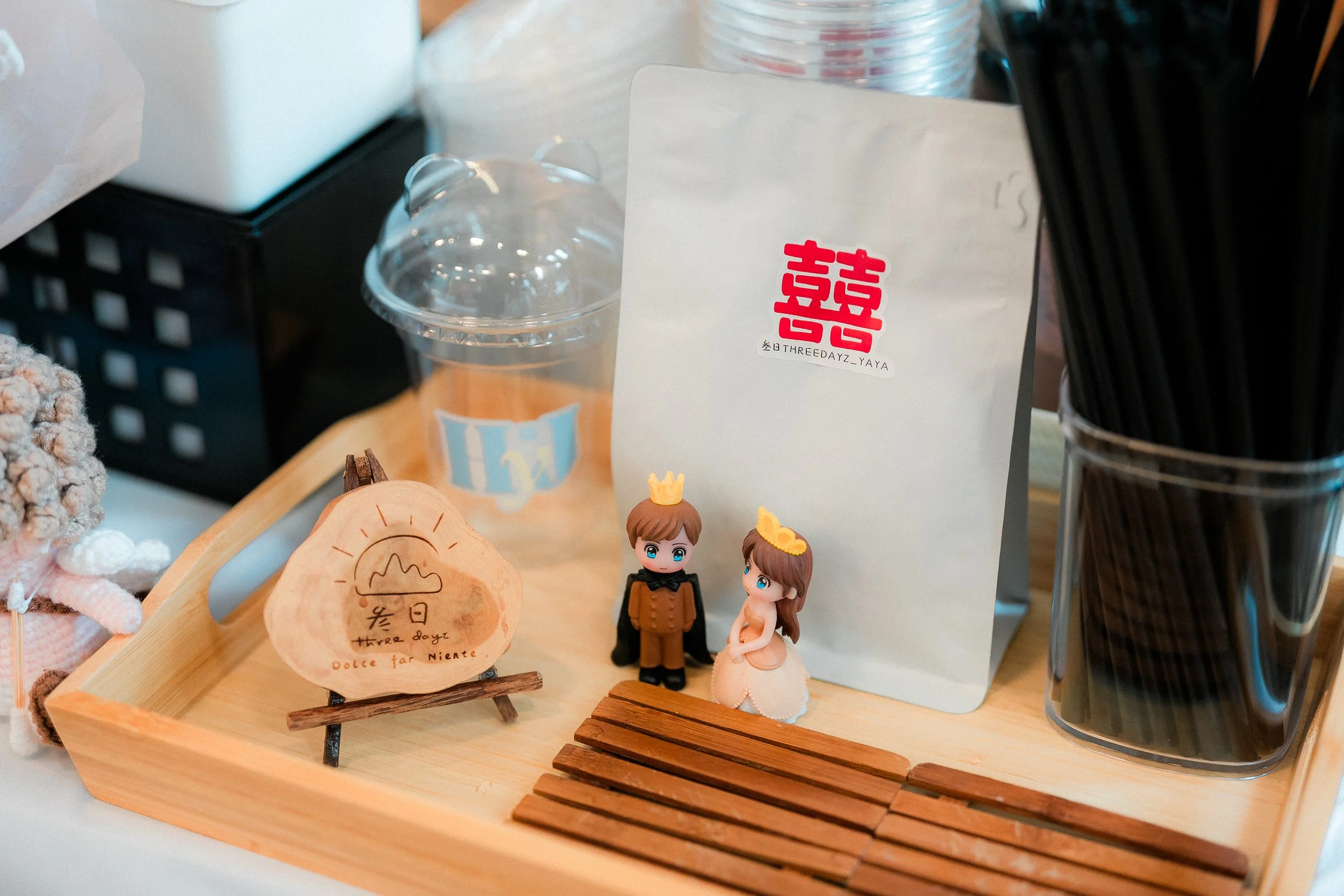 Two small figurines, one of a boy and one of a girl, wearing crowns, are standing on a wooden tray with small wooden planks. There is a paper bag with red and black text behind them, a clear plastic cup with a label, and another container of black st